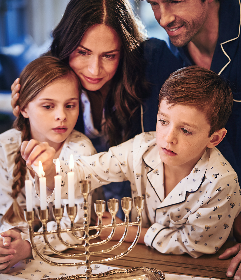 A family wearing pajamas gathers closely, lighting candles on a silver menorah for Hanukkah. The mother hugs her daughter while the son lights a candle, with the father smiling in the background.