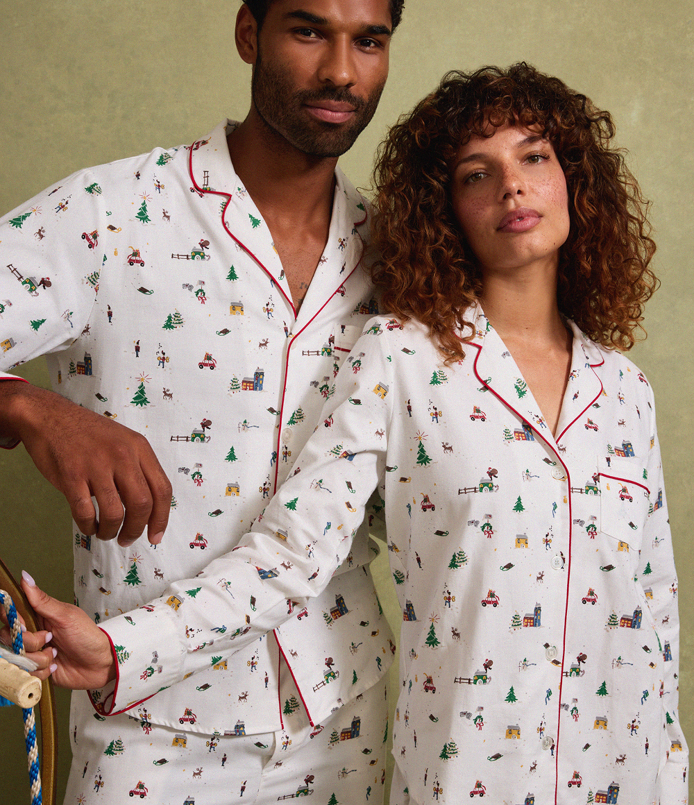 A man and a woman pose together wearing matching white pajamas with festive prints, including trees and tractors. The pajamas have red piping and button-down tops. The background is olive green.