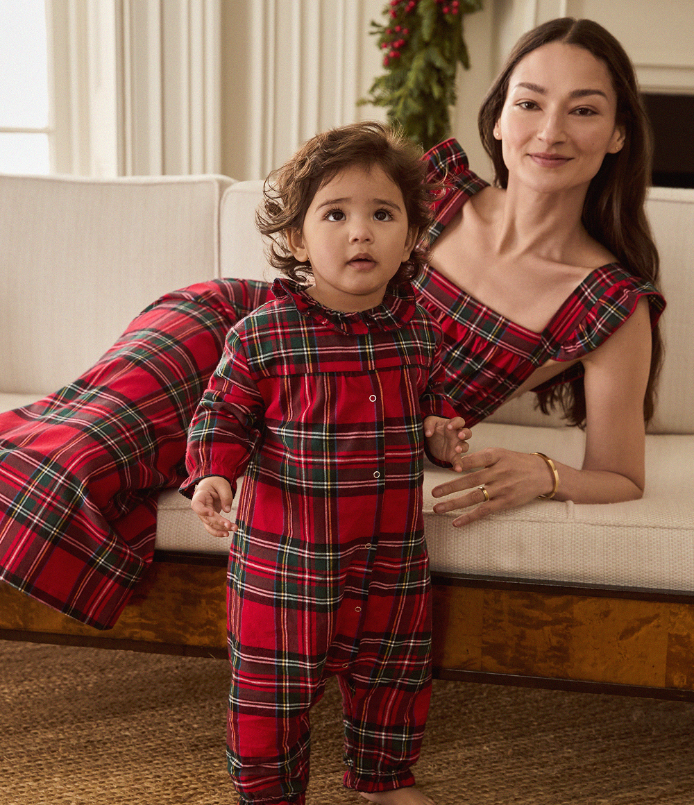 A woman and a young child, both wearing matching red plaid outfits, pose together in a cozy living room. The woman is reclining on a sofa while the child stands in front of her.