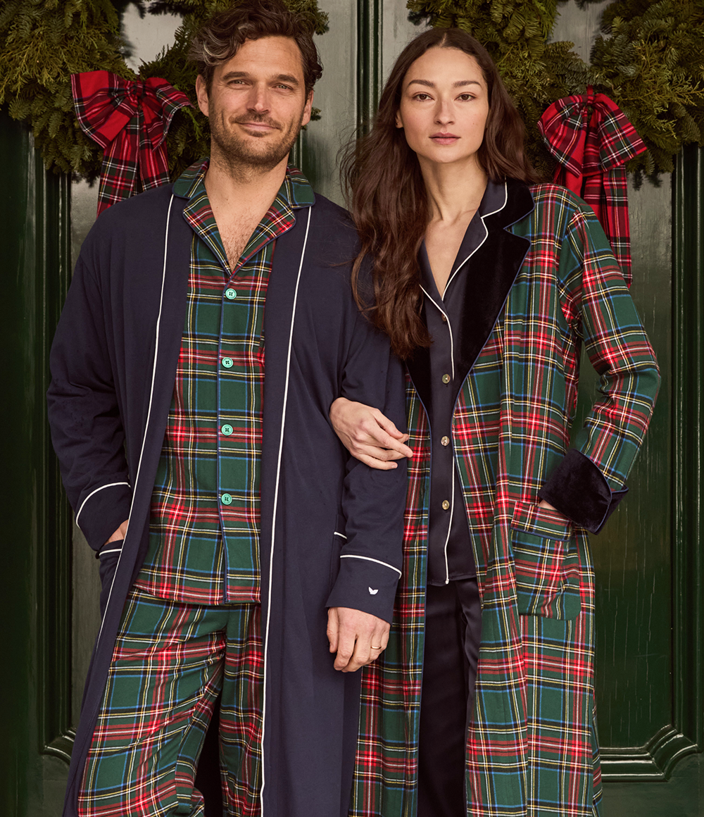 A man and woman stand arm in arm, wearing matching green and red plaid pajamas in front of double doors decorated with festive wreaths and red plaid bows.