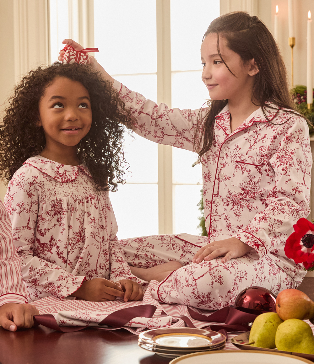 Two young girls in matching floral pajamas sit together indoors. One girl smiles while placing a small red gift box on the other’s head. A table with fruit, a flower, and Christmas ornaments is in the foreground.