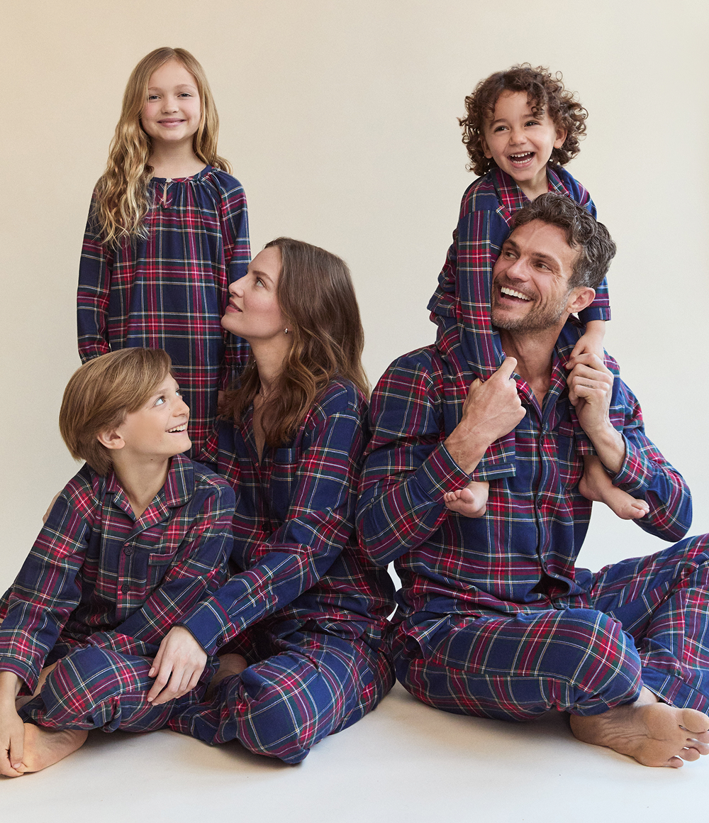 A family of five wearing matching plaid pajamas sits and smiles together. The parents sit on the floor with their three children, one of whom is sitting on the fathers shoulders. Everyone looks happy and relaxed.