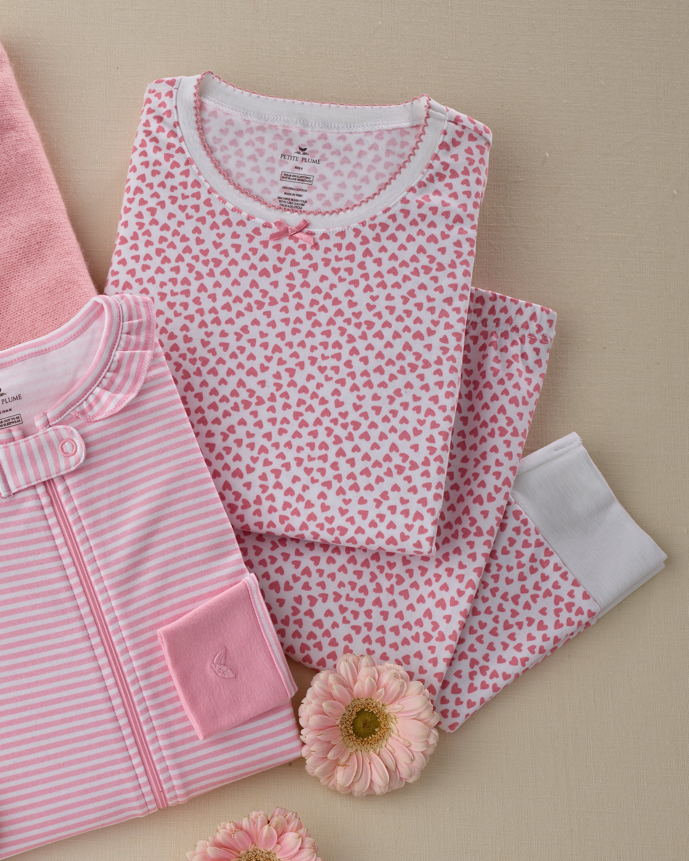 On a beige surface with two pink flowers lie two folded Petite Plume baby outfits. The Kids Pima Snug Fit Pajama Set in Sweethearts features white fabric with pink hearts, while the second is pink with white stripes and a pocket, both offering cozy, luxurious sleepwear for little ones.