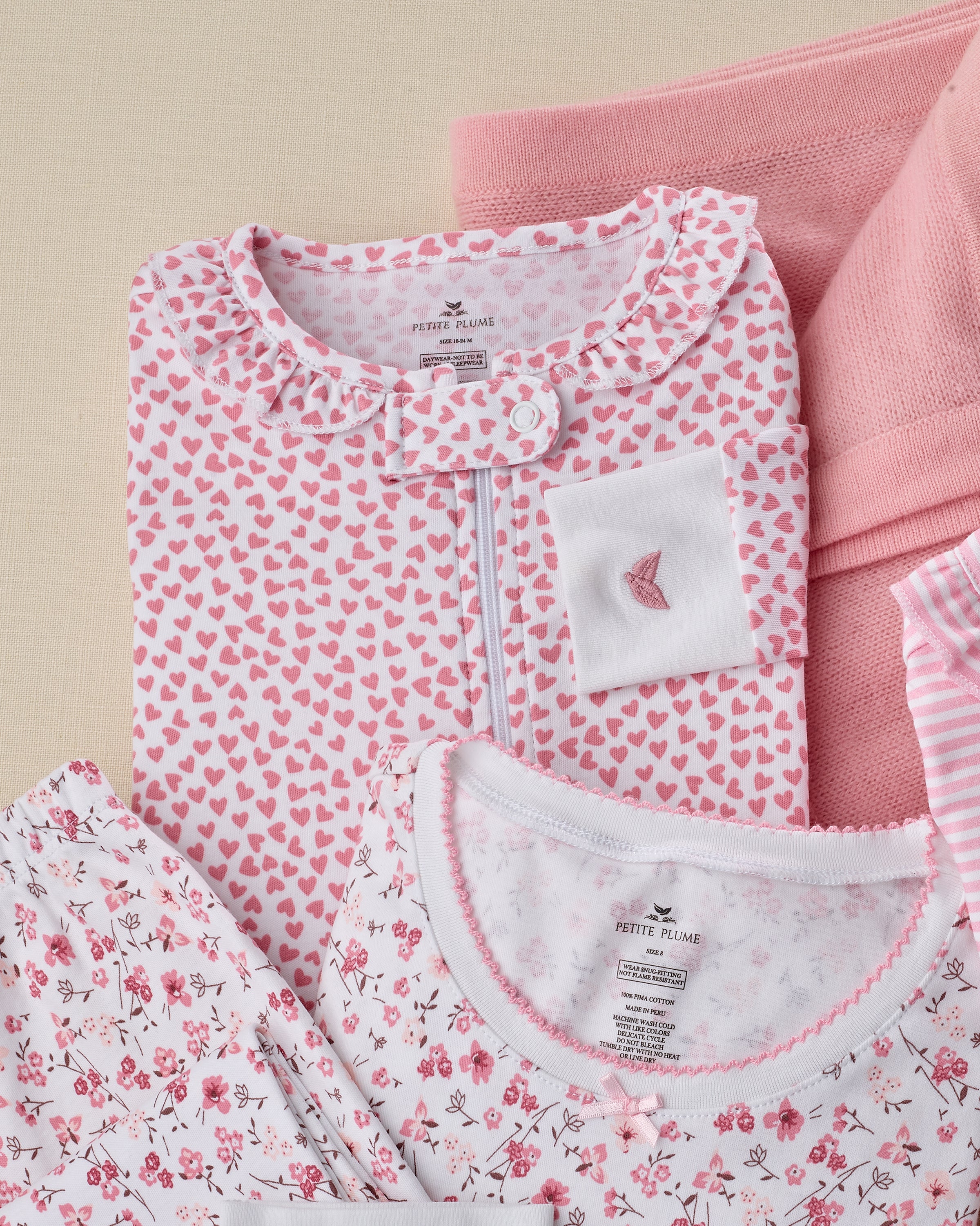 A neatly arranged stack of childrens clothing showcases Petite Plumes Baby Pima Ruffle Collar Romper in Sweethearts with pink and white heart and floral patterns. The set includes a top with a pocket, a striped piece, on a light surface with a soft pink blanket.