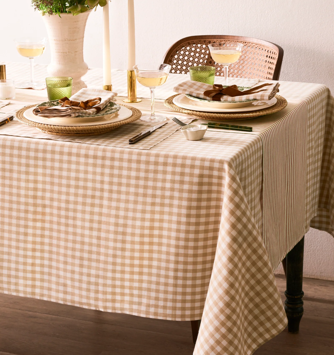 A table set for two with beige and white gingham tablecloth, gold-rimmed plates, green glasses, candles, and wine glasses. Wooden chair and decorative vase with flowers are also visible.
