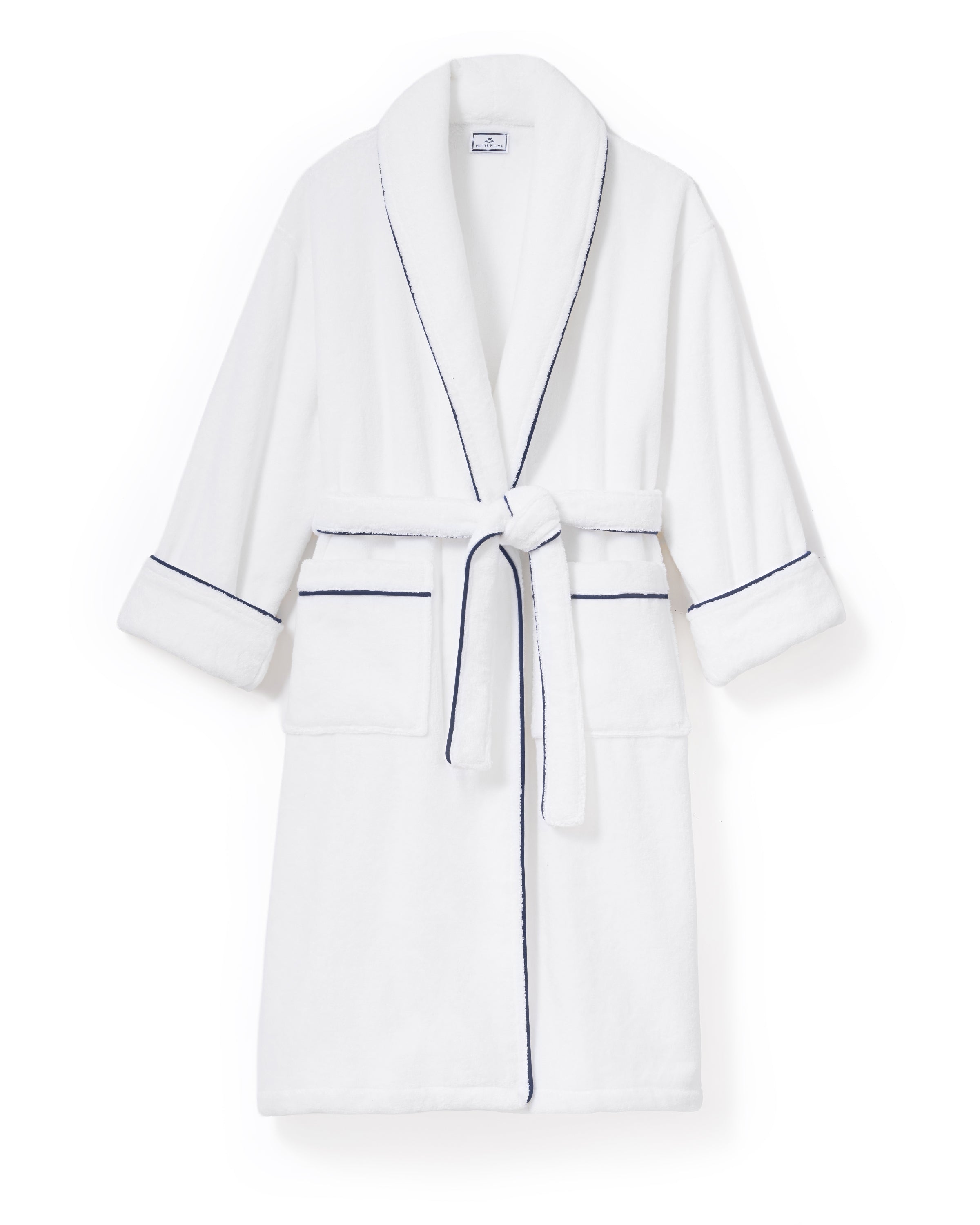 The Unisex Luxe Terry Robe in White with Navy Piping has a shawl collar, front pockets, waist tie, rolled sleeves, and dark piping along the edges.