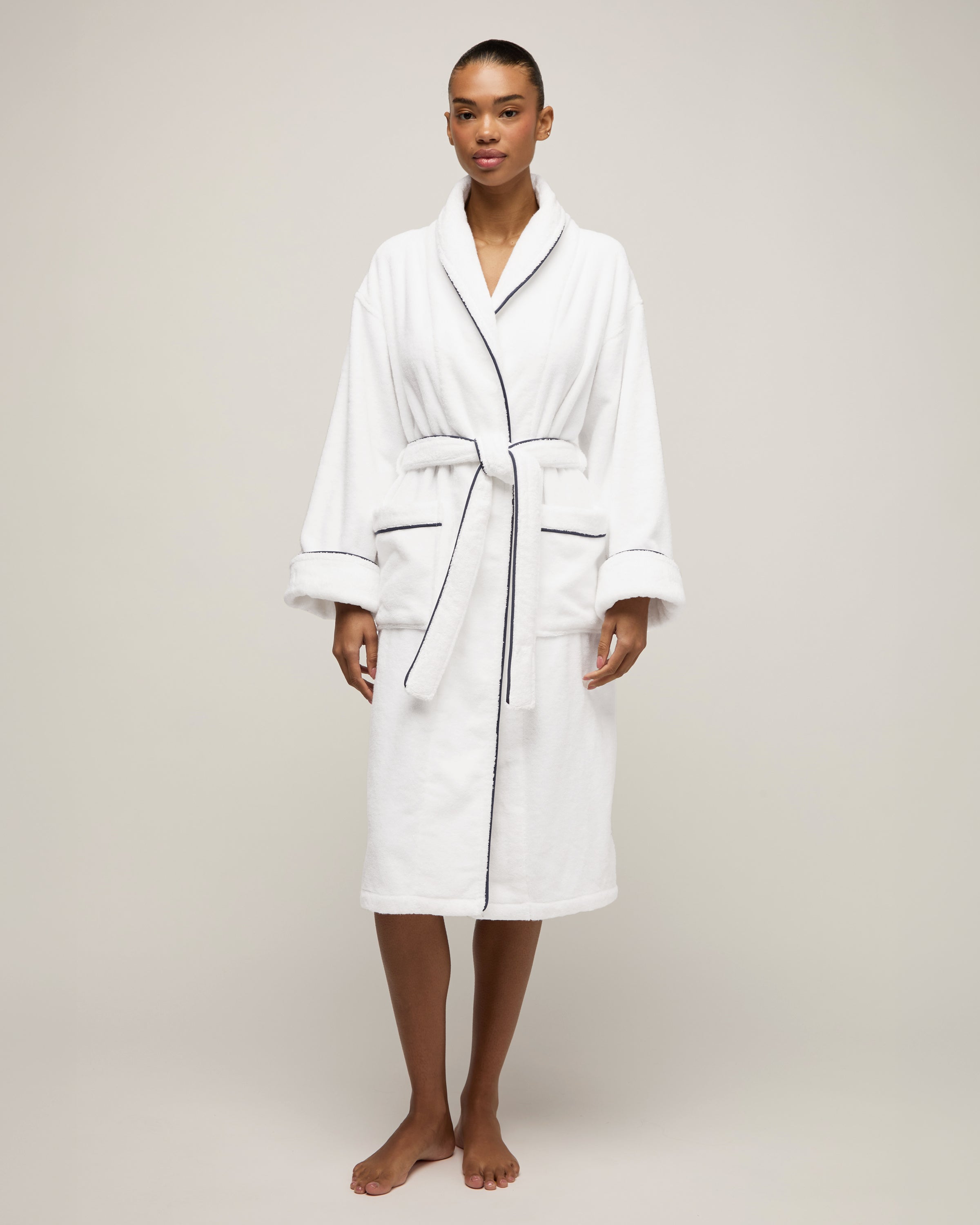 The Unisex Luxe Terry Robe in White with Navy Piping has a shawl collar, front pockets, waist tie, rolled sleeves, and dark piping along the edges.