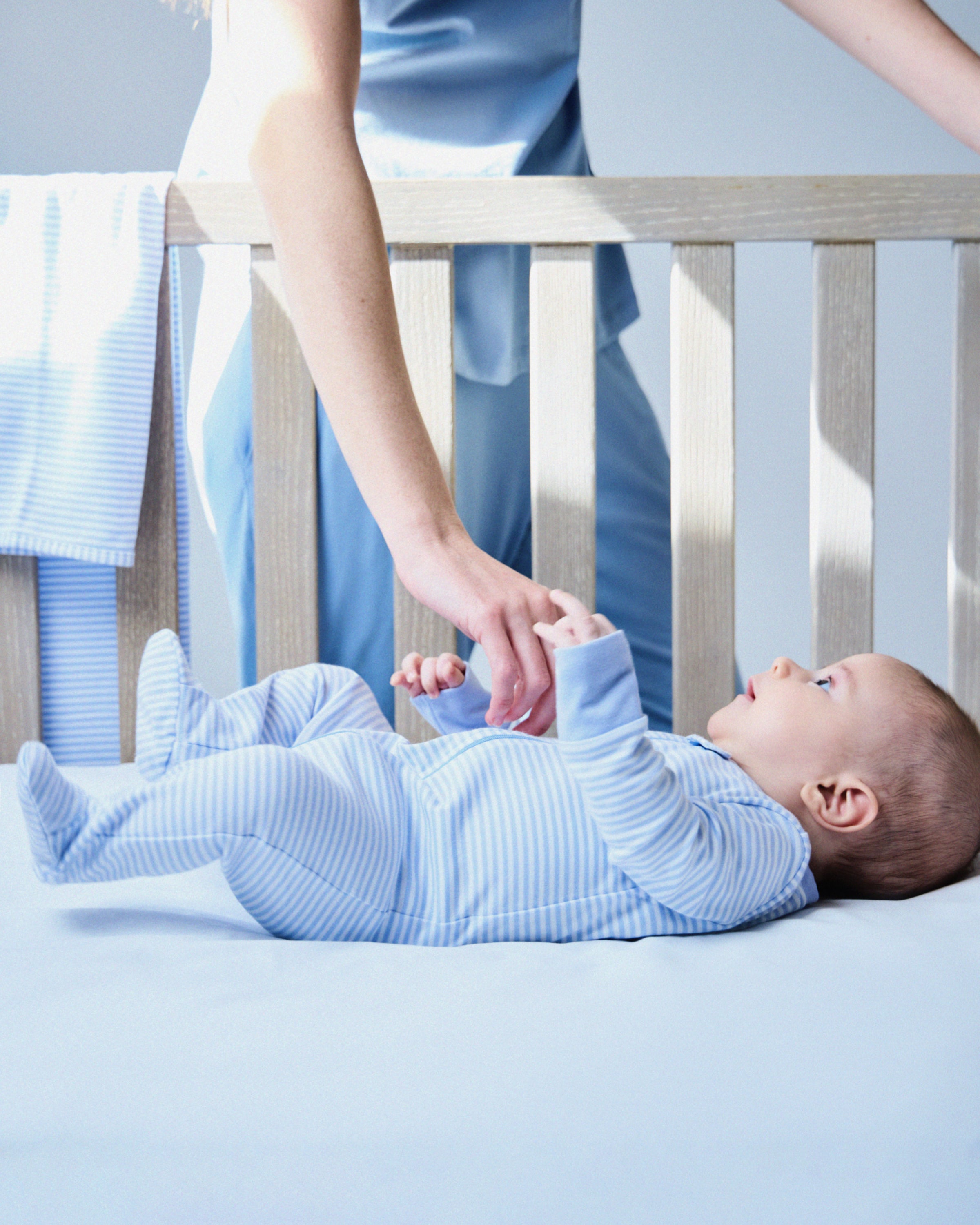 A baby wearing Petite Plumes Babys Pima Romper in Blue Stripes lies in a crib, reaching for an adults hand. The adult, also dressed in blue Pima cotton, leans over the wooden rails draped with a coordinating striped blanket.