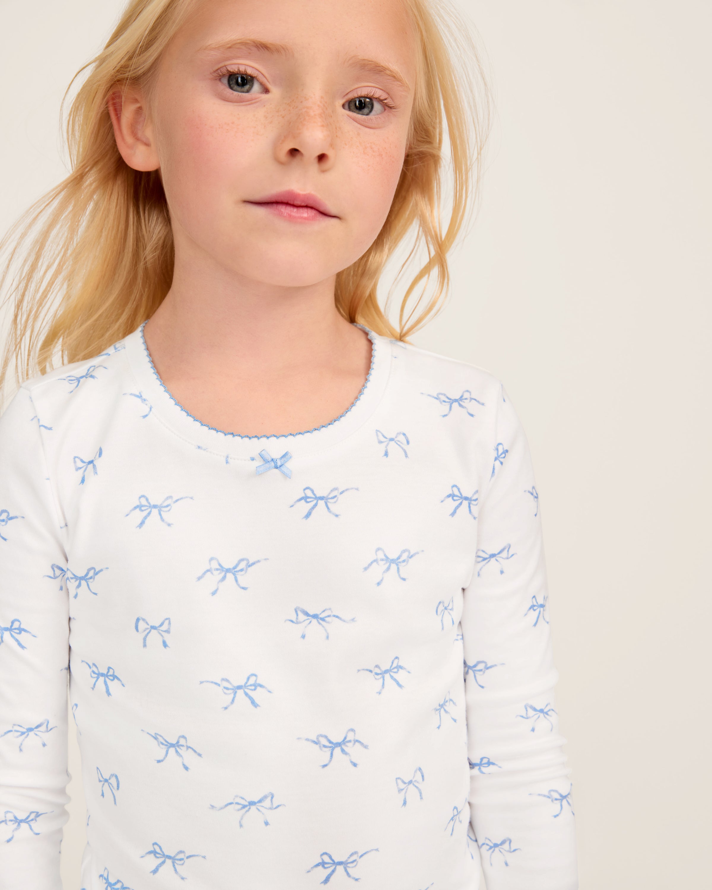 The Kid's Pima Snug Fit Pajama Set in Something Blue Bows features a long-sleeve white top and matching pants with a light blue bow pattern, crew neckline, and soft, comfortable fabric.