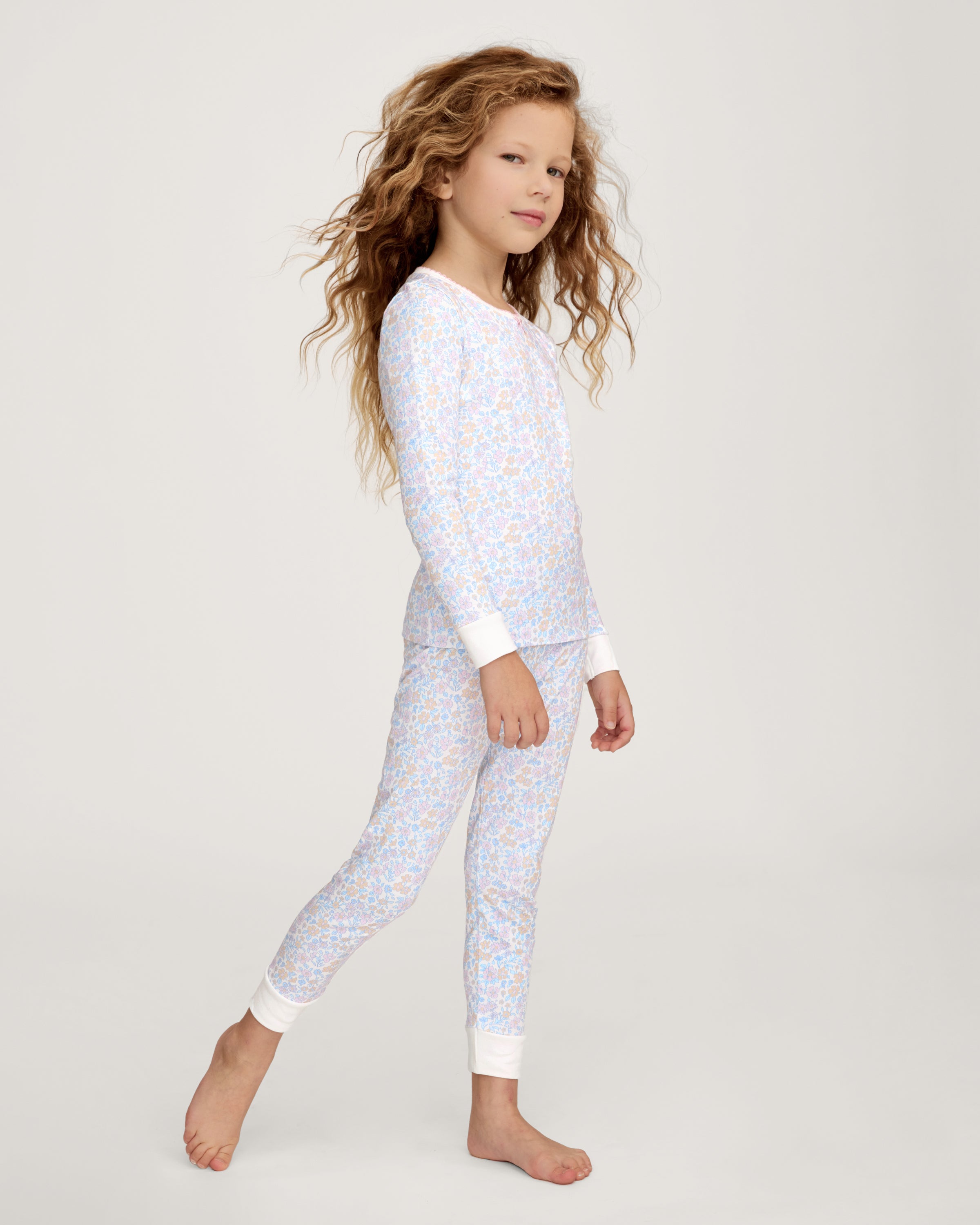 A young girl with long, wavy hair stands barefoot on a plain background, wearing the Kid's Pima Snug Fit Pajama Set in Fleur Blanche. She looks slightly to the side with a relaxed expression.