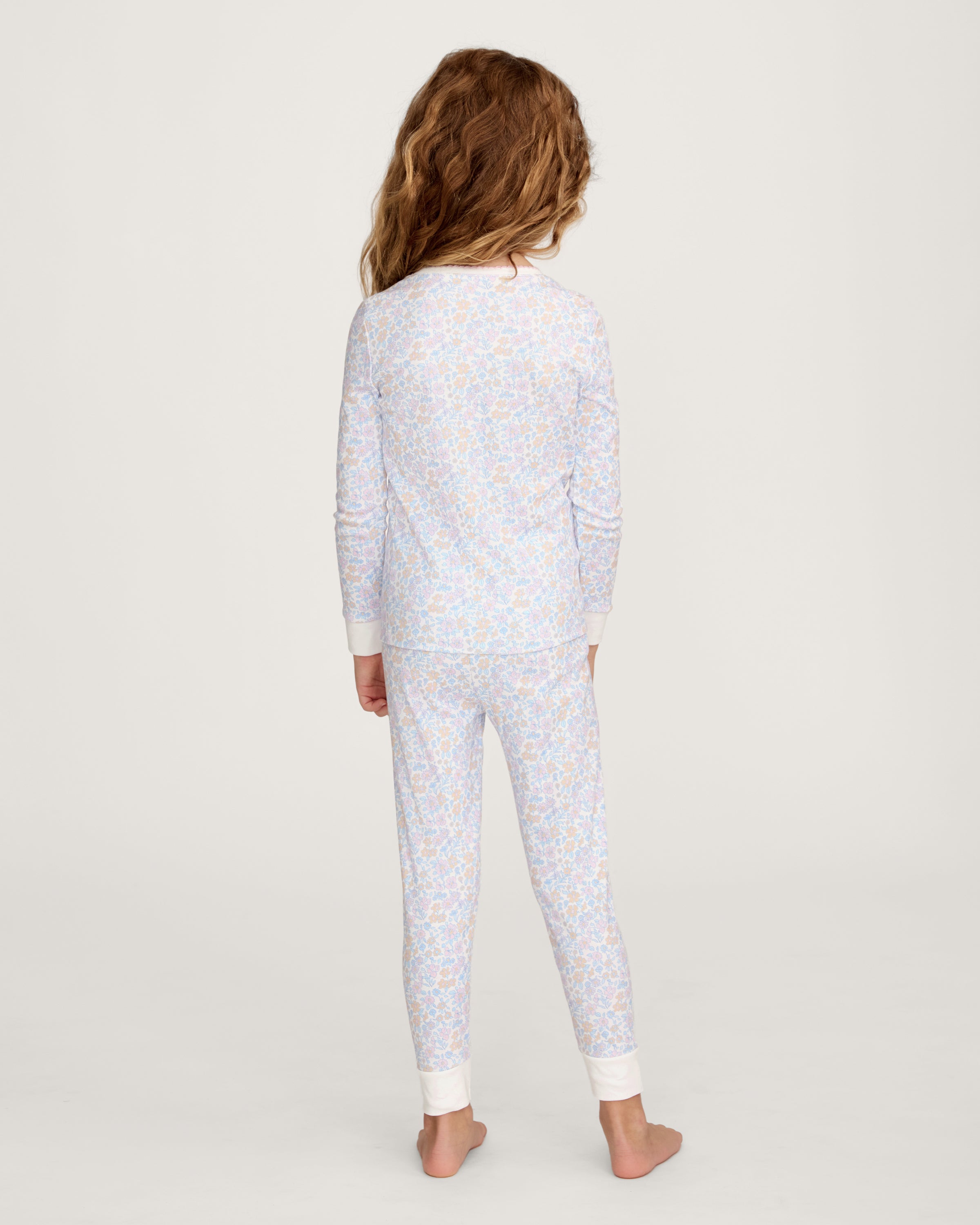 A young girl with long, wavy hair stands barefoot on a plain background, wearing the Kid's Pima Snug Fit Pajama Set in Fleur Blanche. She looks slightly to the side with a relaxed expression.