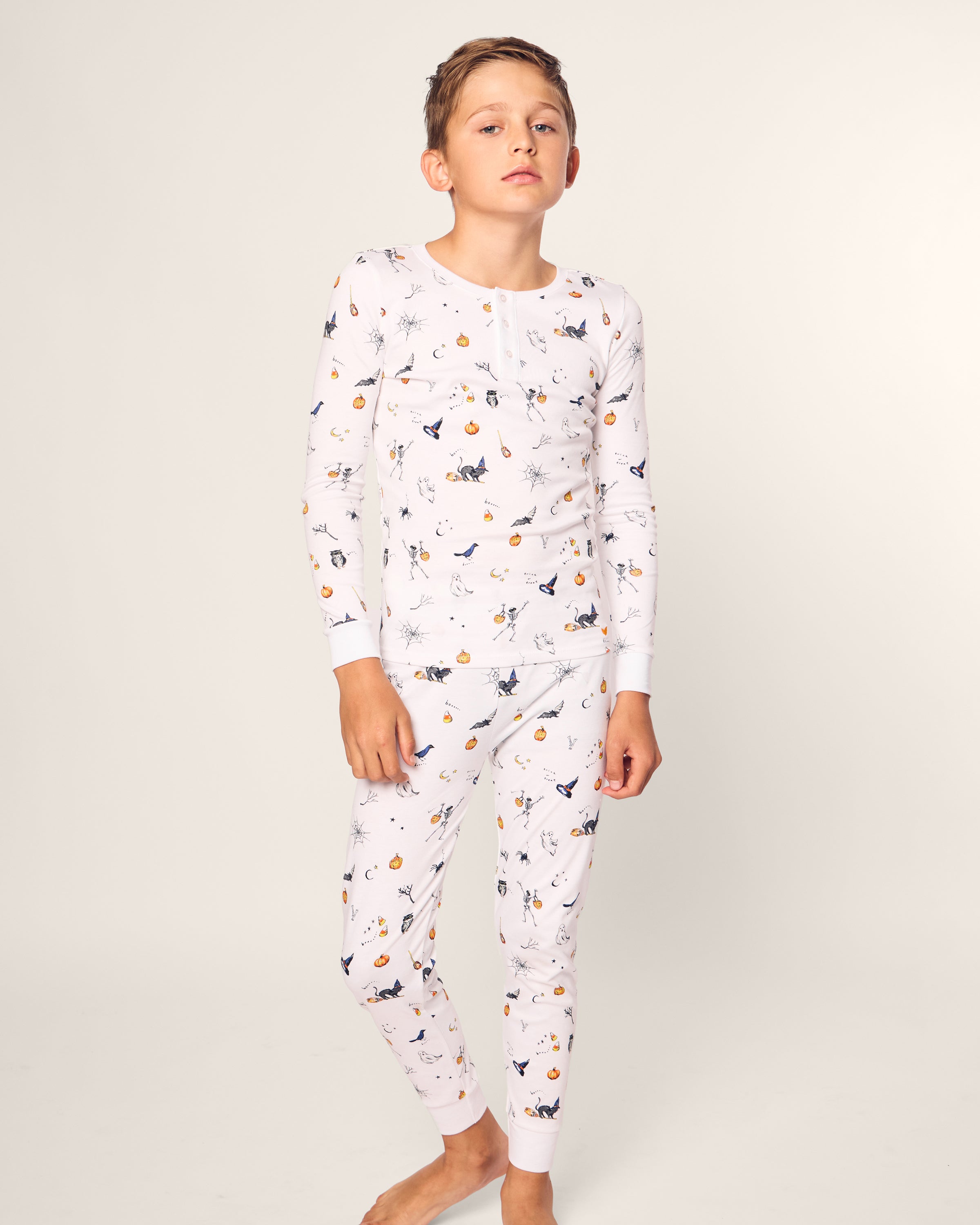 A young person stands barefoot in a relaxed pose, wearing the Petite Plume Kids Pima Snug Fit Pajama Set in Trick or Treat, crafted from luxurious Pima cotton with whimsical animal and nature patterns. The background is plain light gray.