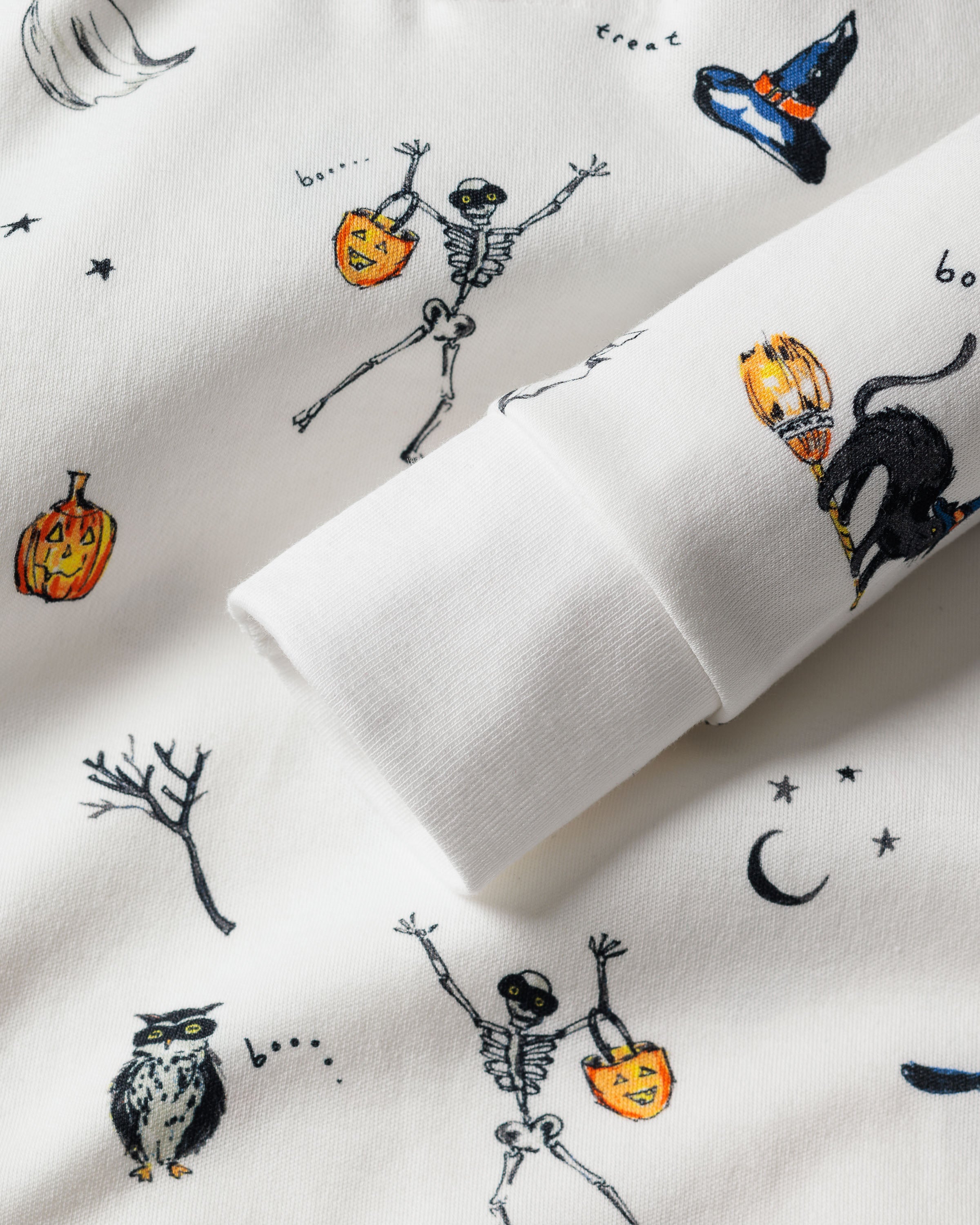 Close-up of the Kids Pima Snug Fit Pajama Set in Trick or Treat from Petite Plume, showcasing Halloween-themed illustrations like skeletons, jack-o-lanterns, and more on a white fabric. The long sleeve top features playful designs on soft Pima cotton.