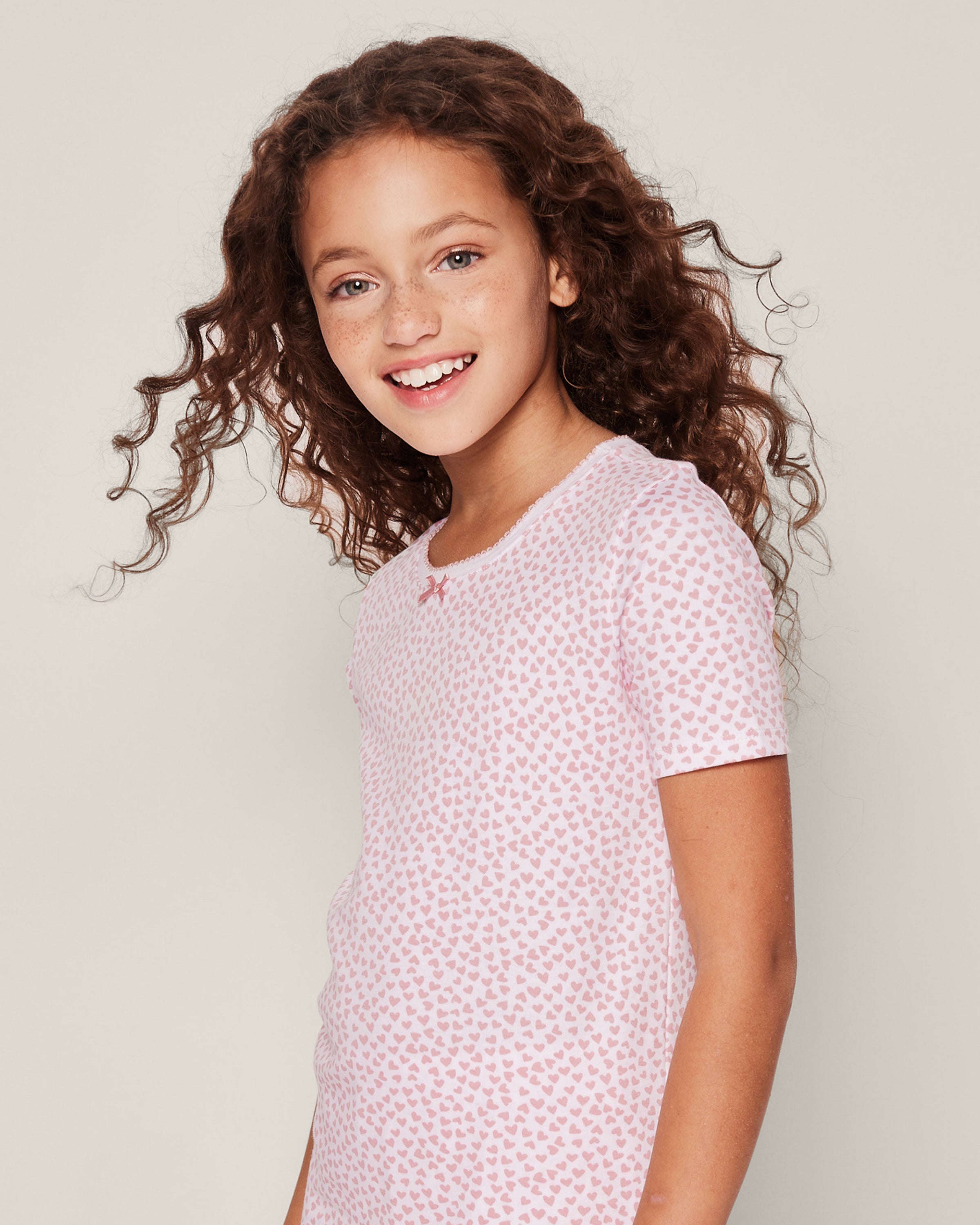 A young girl with long, curly hair smiles while wearing the Kids Pima Snug Fit Pajama Short Set in Sweethearts by Petite Plume. The pink and white patterned luxury sleepwear features cozy long sleeves and shorts, adorned with small heart designs, against a plain background.