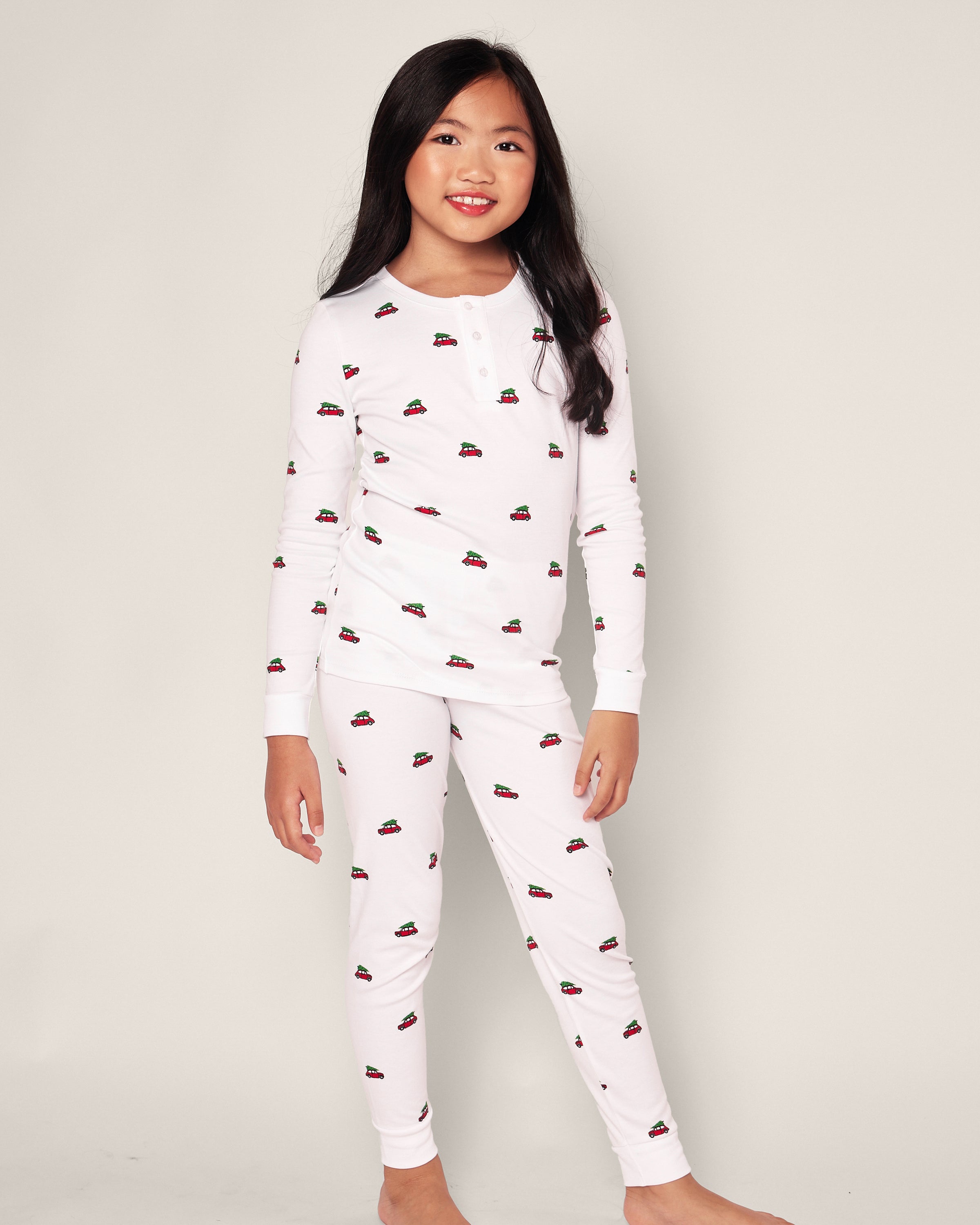 A young girl stands barefoot in Petite Plumes Kids Pima Snug Fit Pajama Set in Holiday Journey. Her long dark hair flows freely as she smiles against a plain light background, showcasing the pajamas white long sleeve top and pant set adorned with small car illustrations.
