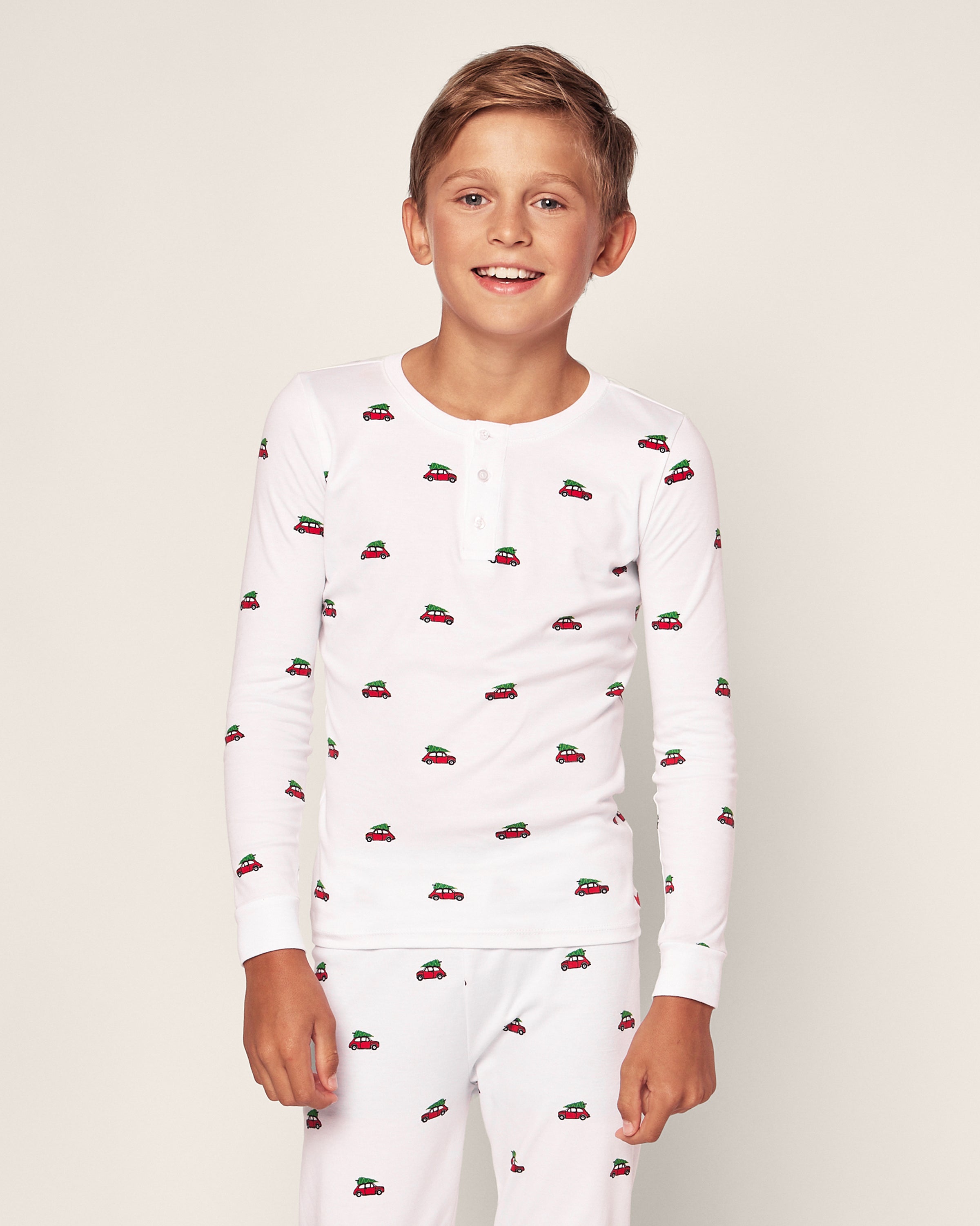 A smiling boy wears Petite Plumes Kids Pima Snug Fit Pajama Set in Holiday Journey, featuring a pattern of small red cars on white, made from soft pima cotton. He stands against a plain light background in cozy long sleeve tops and pants with neck buttons.