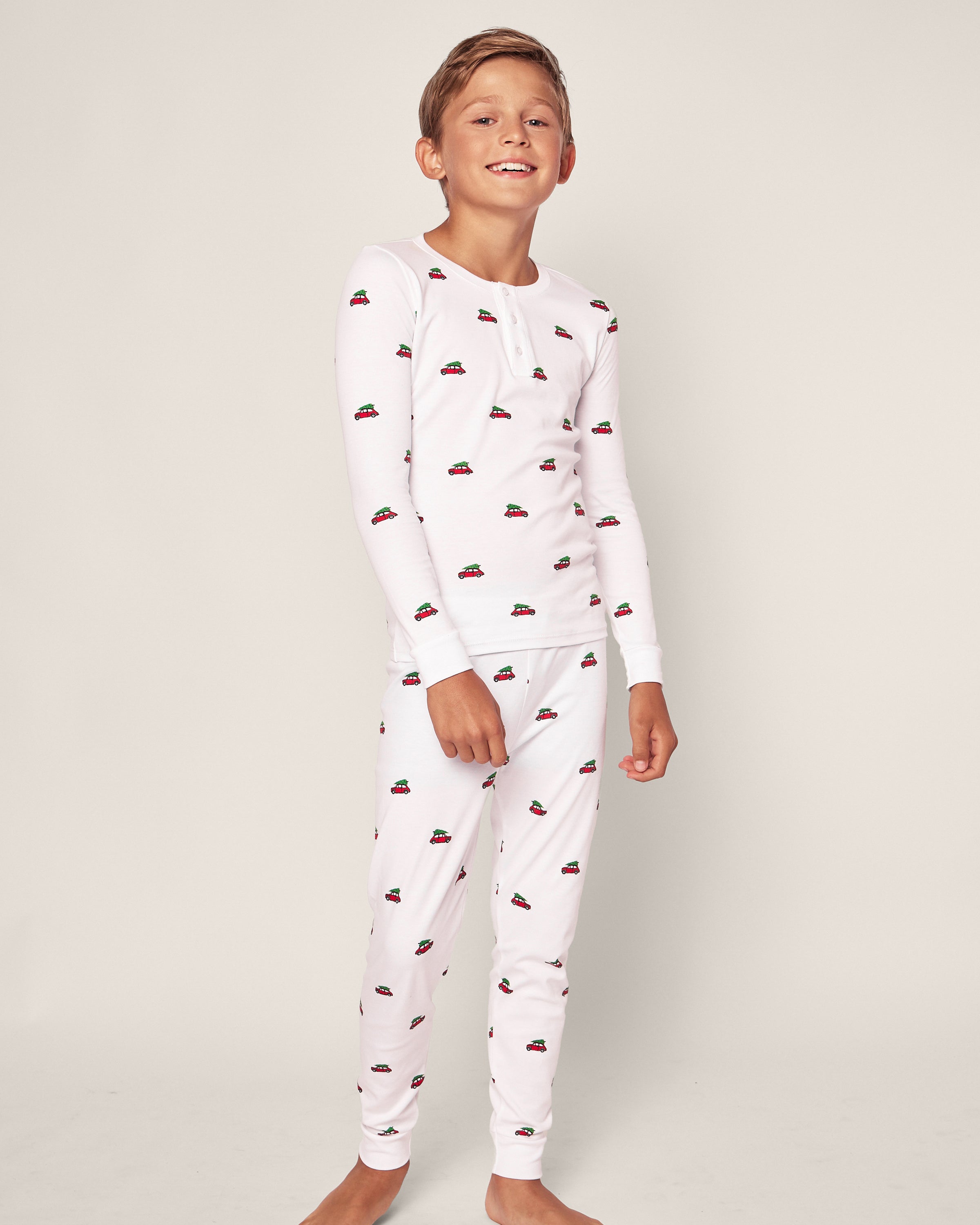 A boy grins widely, dressed in the Kids Pima Snug Fit Pajama Set in Holiday Journey by Petite Plume. The cozy white pajamas are adorned with small red car patterns and crafted from soft Pima Cotton, embodying classic sleepwear charm against a plain light background.