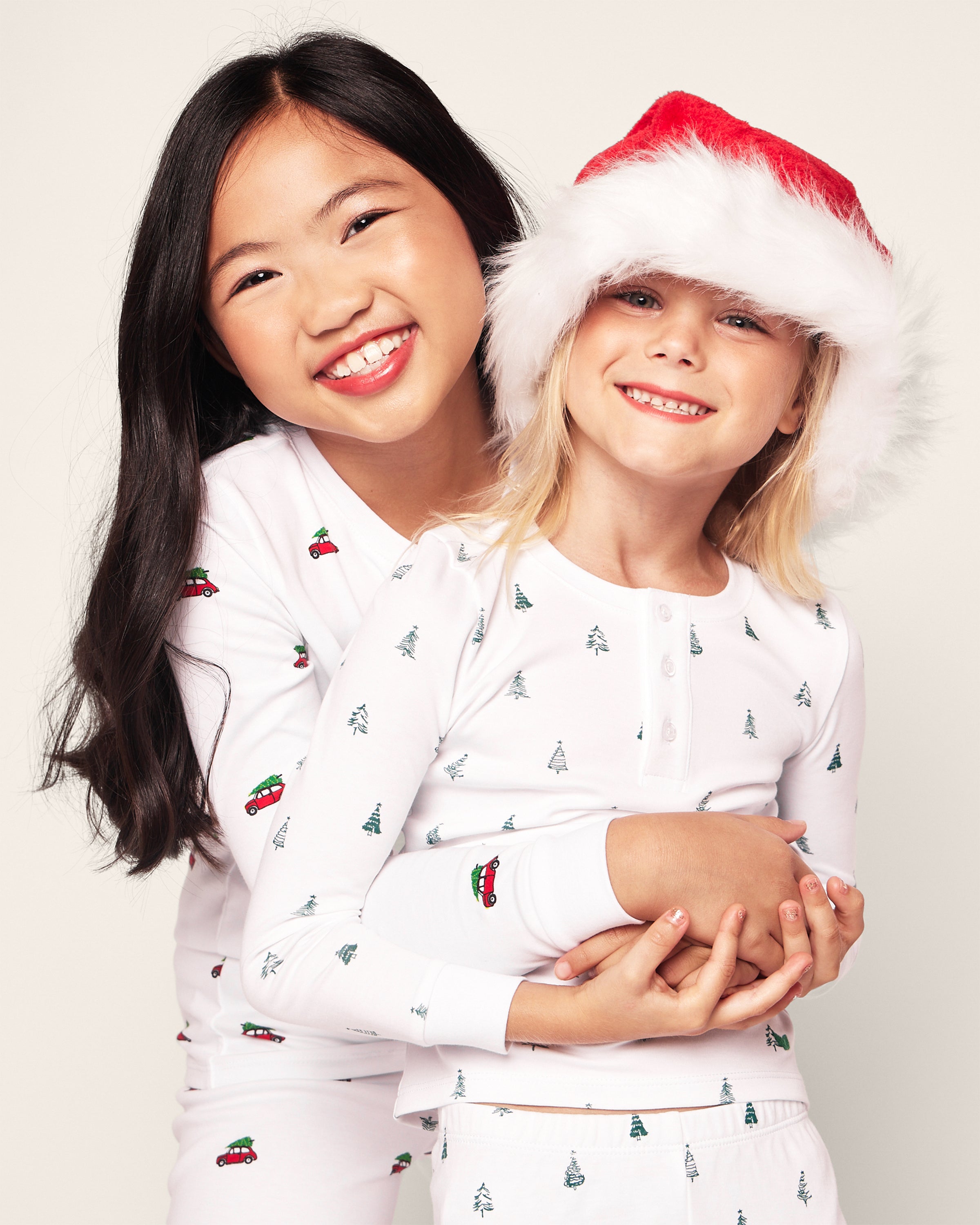 Two children smiling and hugging. The child on the left, with long dark hair, is in Petite Plumes Kids Pima Snug Fit Pajama Set in Holiday Journey style. The child on the right has light hair at shoulder-length, wearing matching pajamas with a Santa hat.