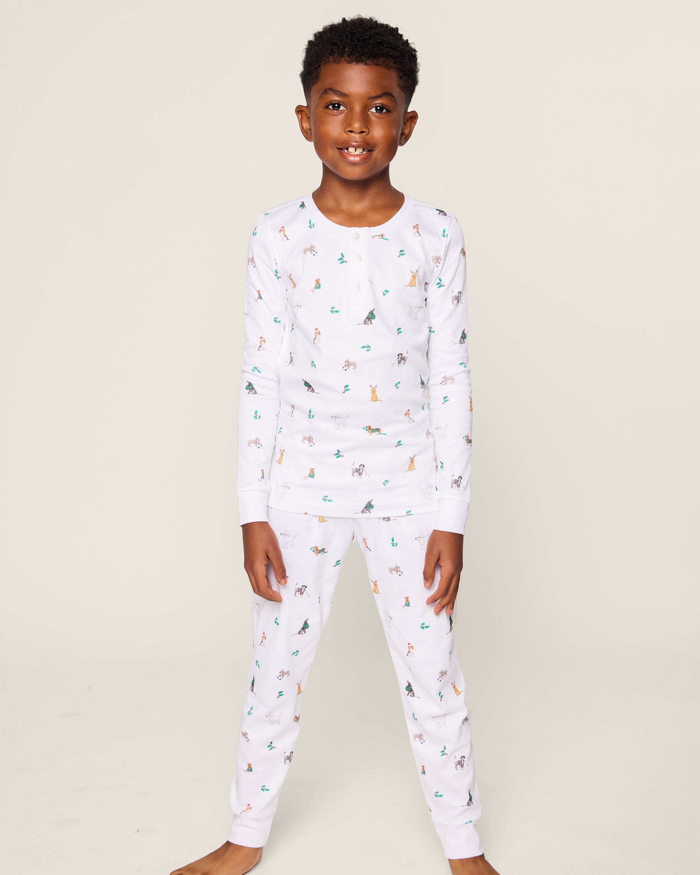A young boy smiles, wearing the Petite Plume Kids Pima Snug Fit Pajama Set in Jingle Paws. This luxurious sleepwear features colorful small animal prints and provides comfort with its long sleeve top and pants as he stands barefoot against a plain background.