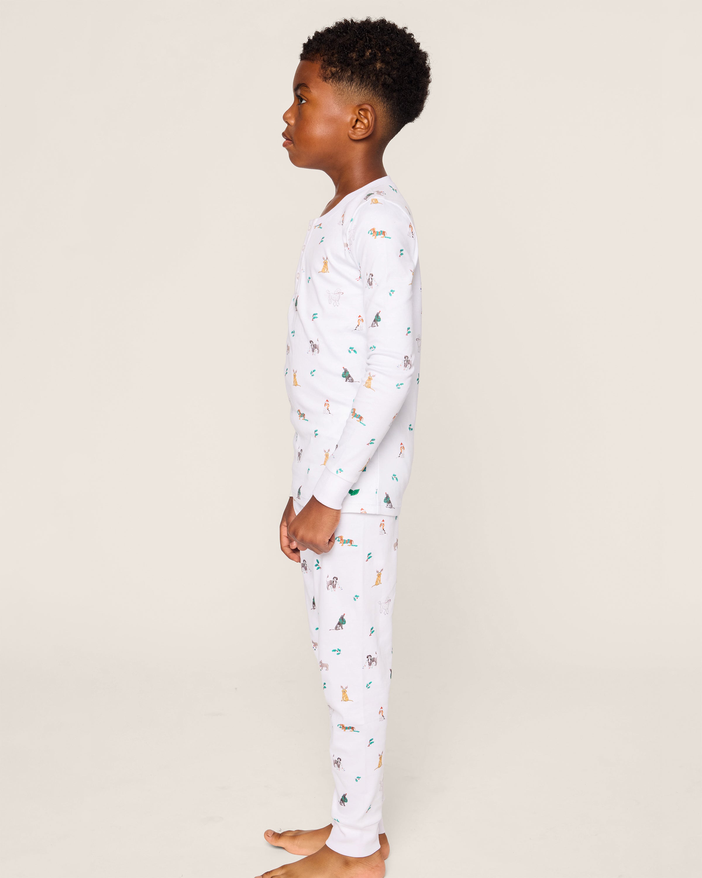 A child stands barefoot against a plain background, wearing Petite Plumes luxurious Kids Pima Snug Fit Pajama Set in Jingle Paws, featuring small colorful animal prints.