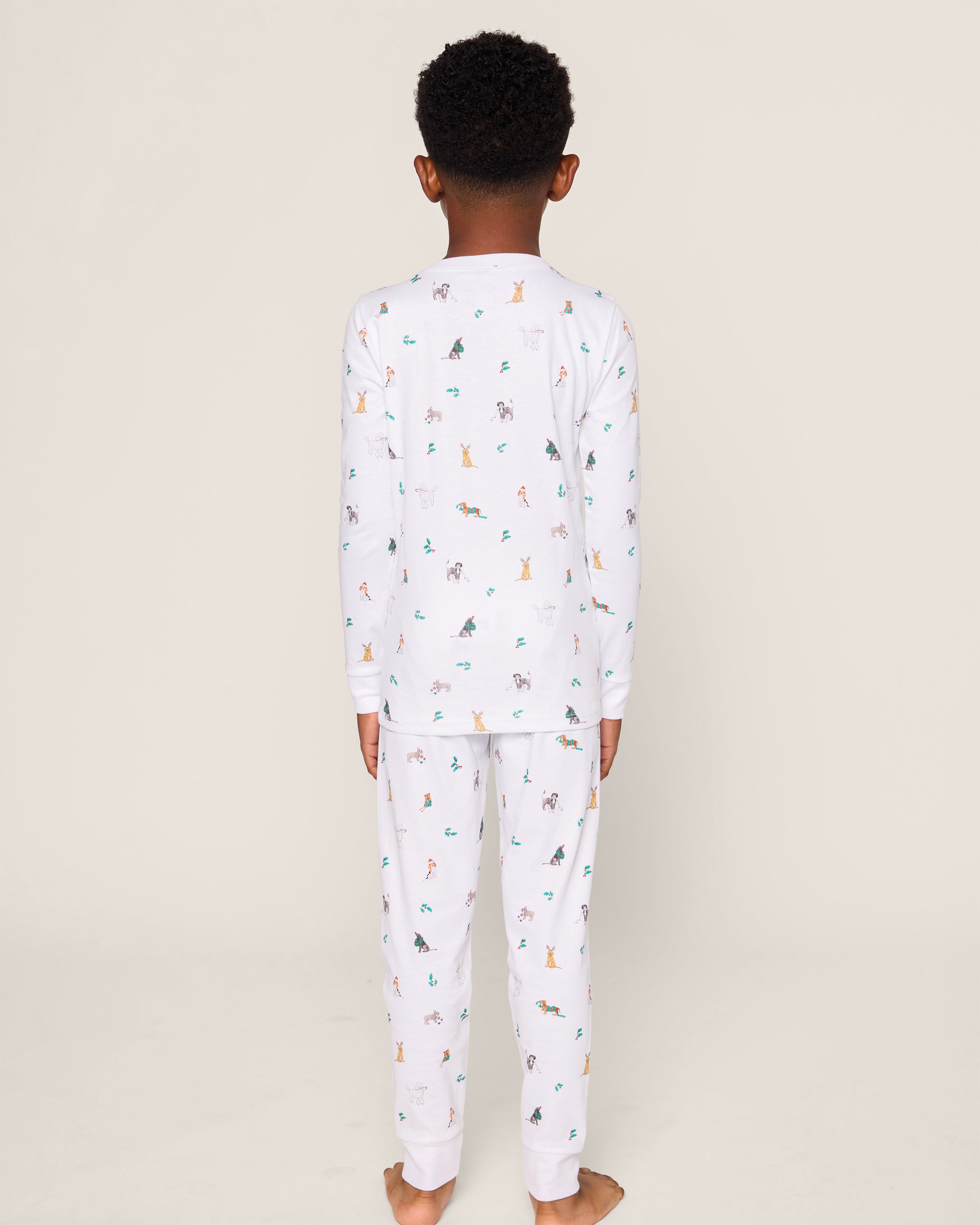 A child stands barefoot on a neutral backdrop, wearing Petite Plumes luxurious Kids Pima Snug Fit Pajama Set in Jingle Paws. The set features soft Pima cotton with colorful cartoon animal prints and long sleeves for added comfort.