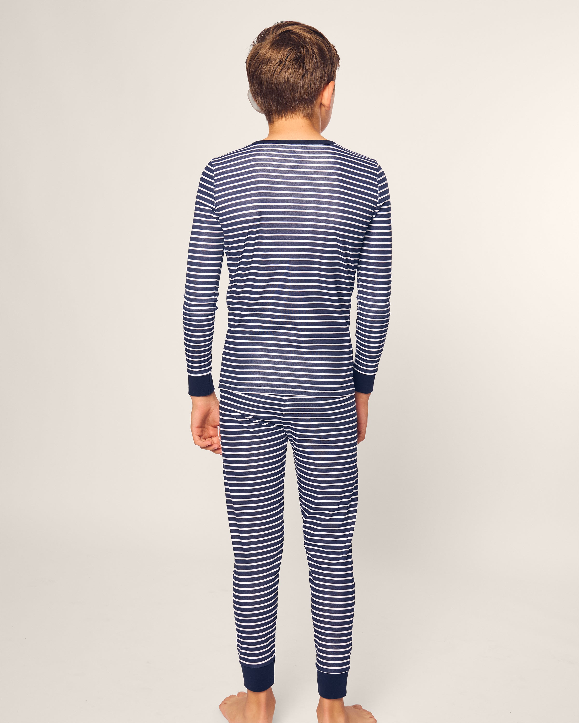 A child stands barefoot, facing away, in a Petite Plume Kids Pima Snug Fit Pajama Set in Navy Stripe. The sleepwears soft texture complements the simple, light-colored background.