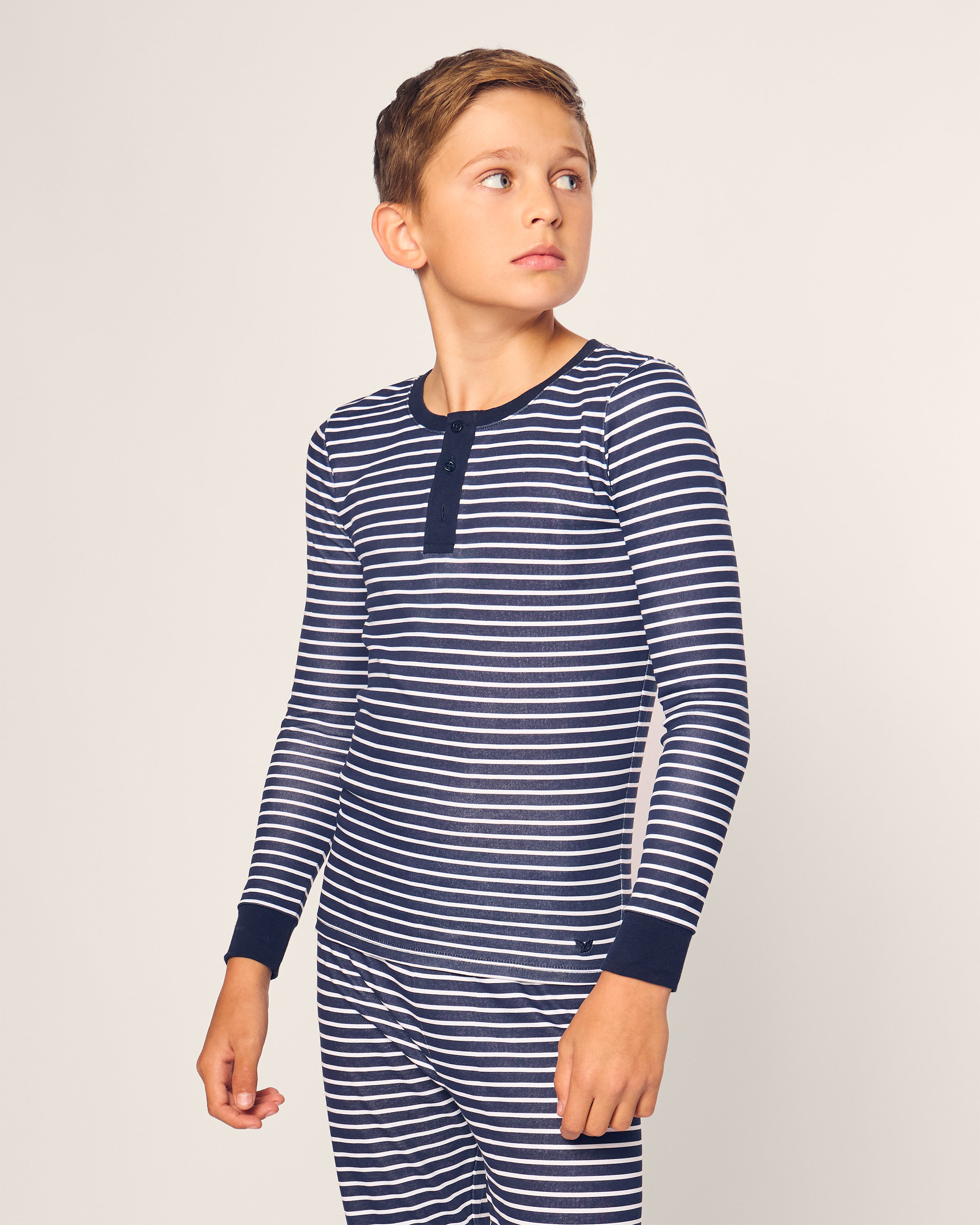 A young boy in the Petite Plume Kids Pima Snug Fit Pajama Set in Navy Stripe stands against a plain background, looking to his right. The Pima cotton top features a buttoned collar for added style.