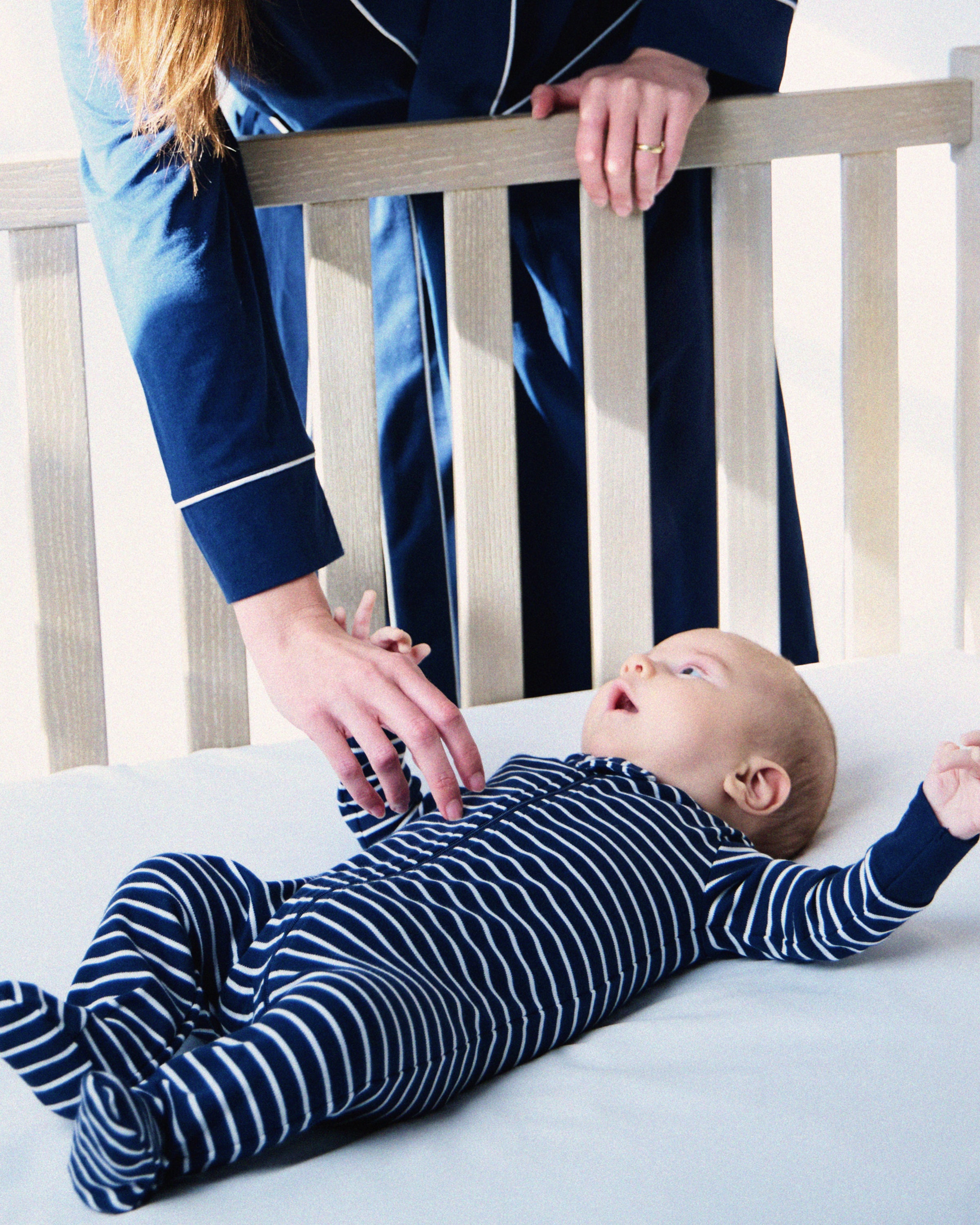 A baby in a navy striped Pima romper from Petite Plume lies in a crib, reaching for a person in a dark blue outfit, who gently touches the babys hand.