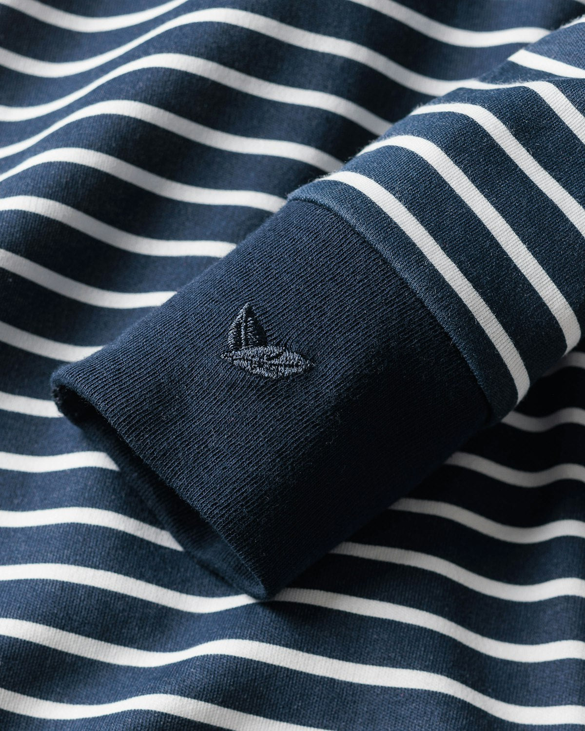 The sleeve of Petite Plumes Babys Pima Romper in Navy Stripe features a close-up of soft Pima cotton, decorated with white horizontal stripes and an embroidered logo of two overlapping triangles in dark blue thread, enhancing the vibrant colors across the collection.