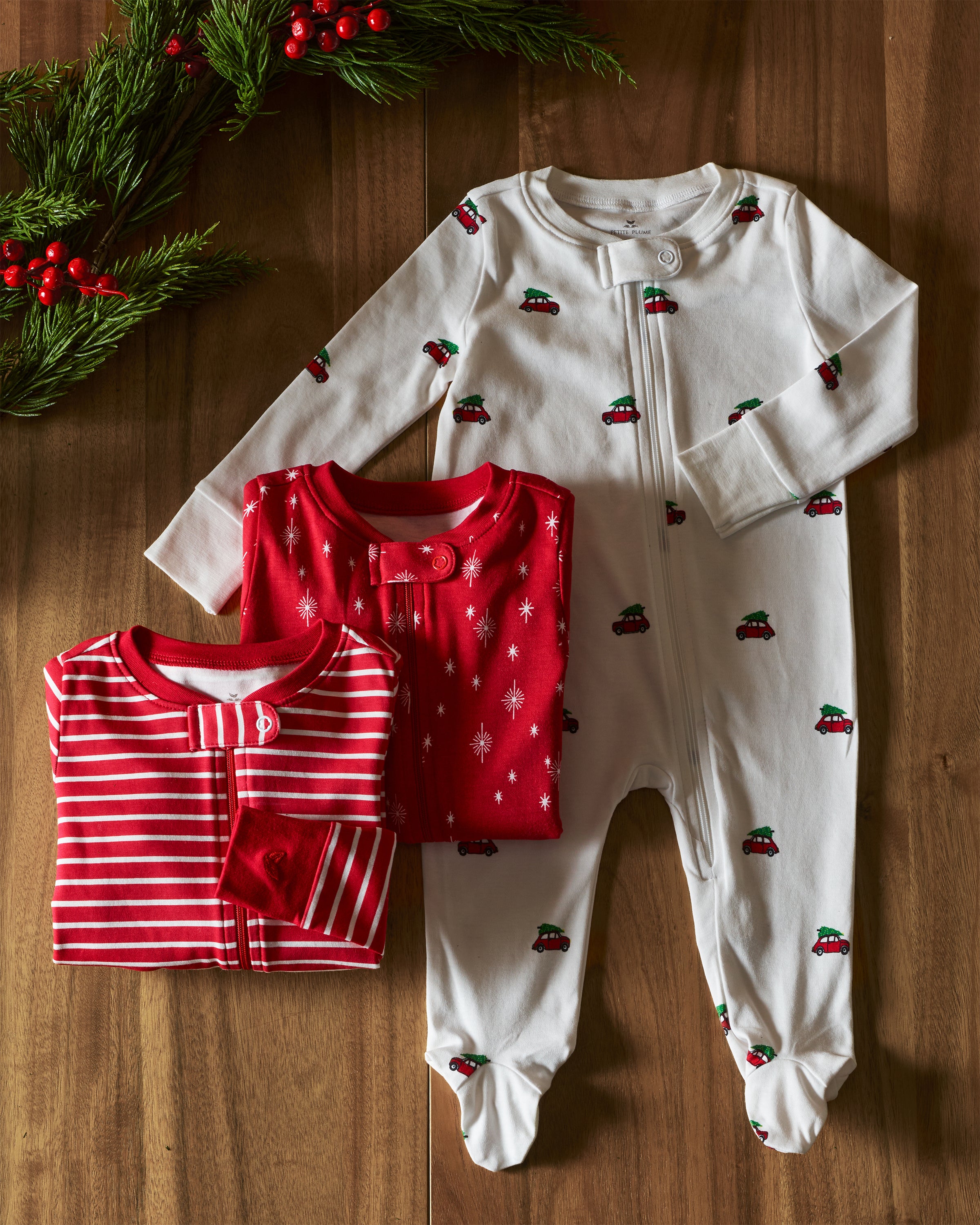 Three ultra-breathable Petite Plume rompers on wood: one white with red car patterns, one red with snowflakes, and another is Babys Pima Romper in Red Stripe. Pine branches and red berries in the corner add a festive touch to these vibrant colors.