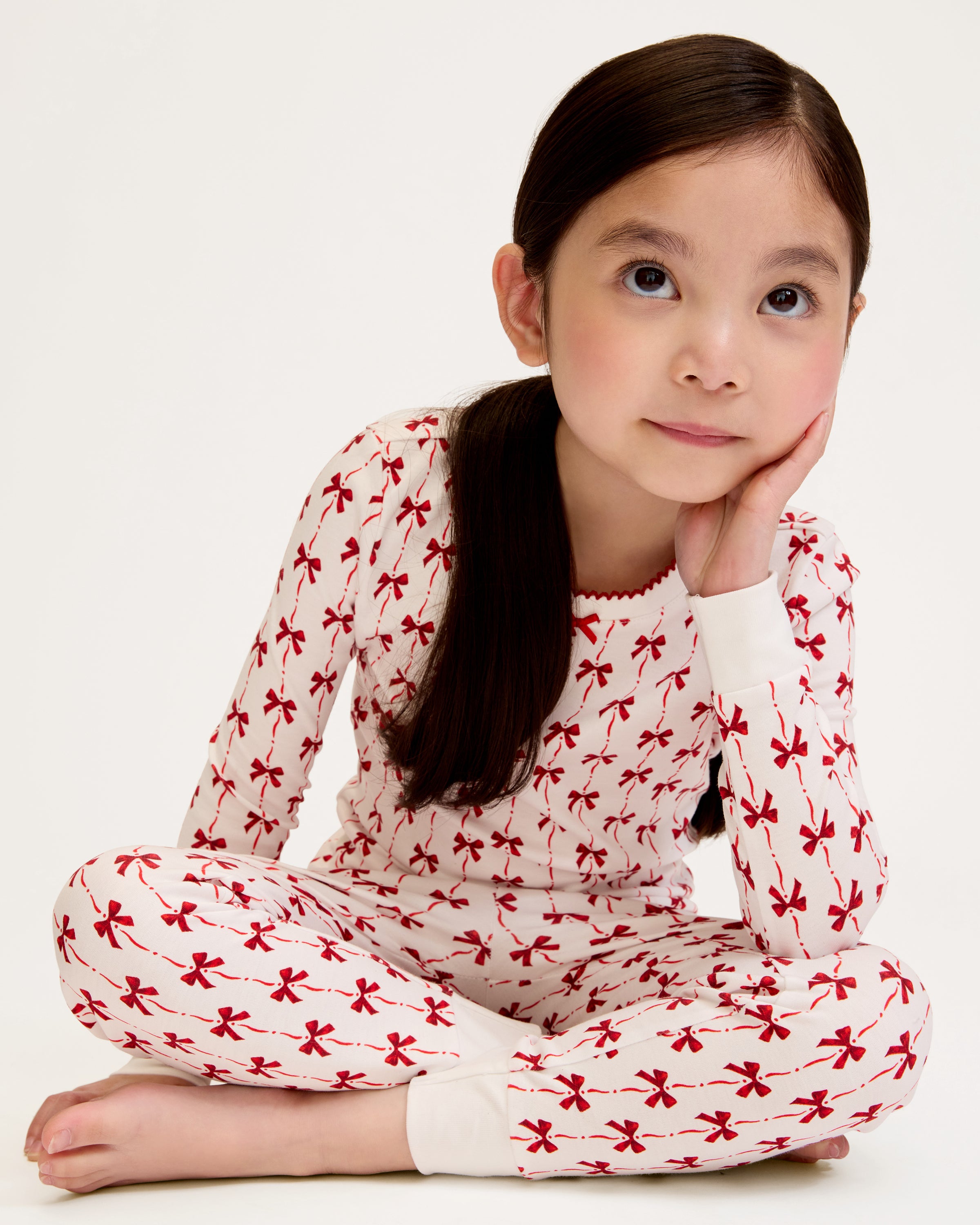 The Kid's Pima Snug Fit Pajama Set in Blissful Bows features a white two-piece design with long sleeves and pants, decorated with a repeating small red bow pattern and finished with white cuffs on the sleeves and pant legs.