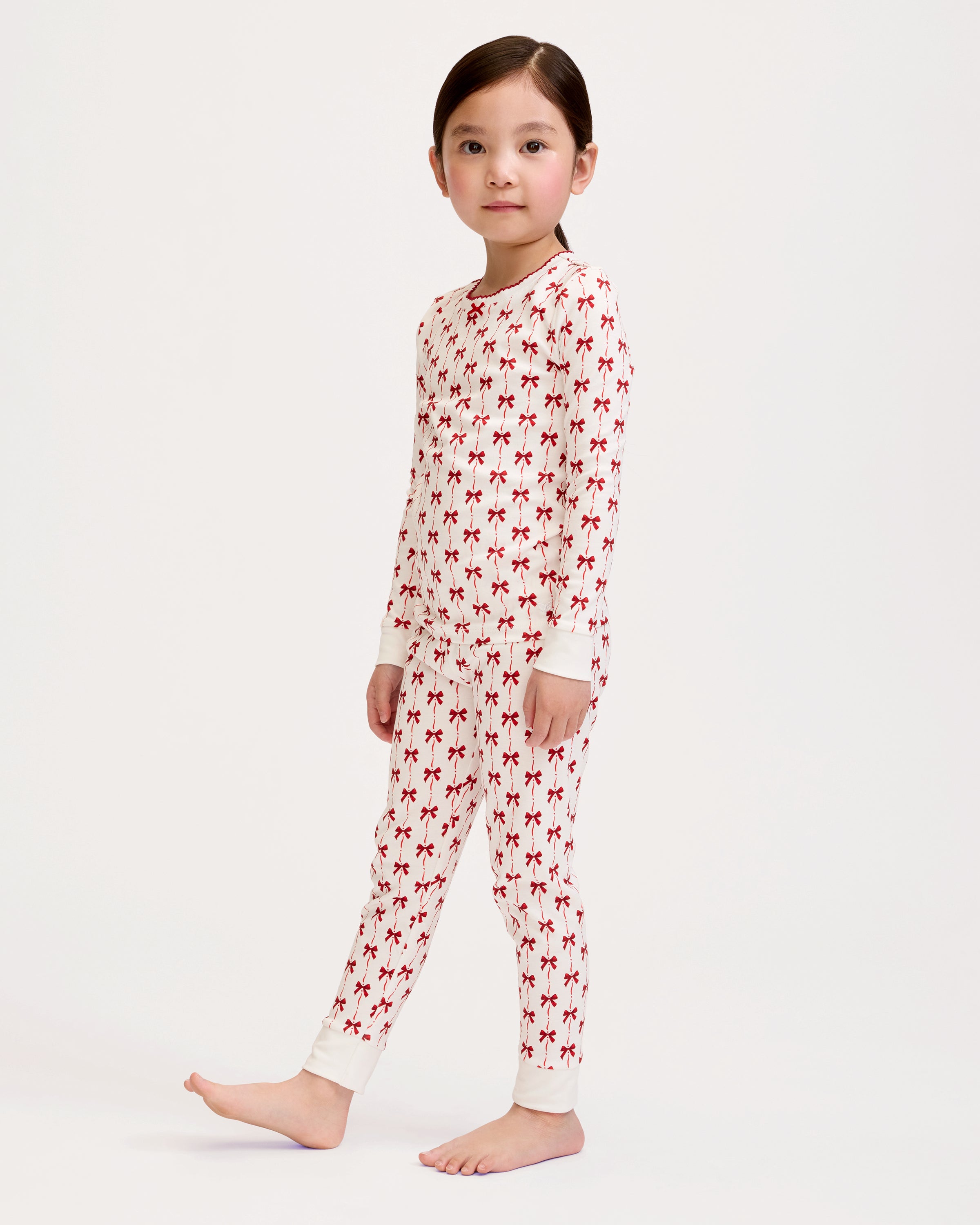 The Kid's Pima Snug Fit Pajama Set in Blissful Bows features a white two-piece design with long sleeves and pants, decorated with a repeating small red bow pattern and finished with white cuffs on the sleeves and pant legs.