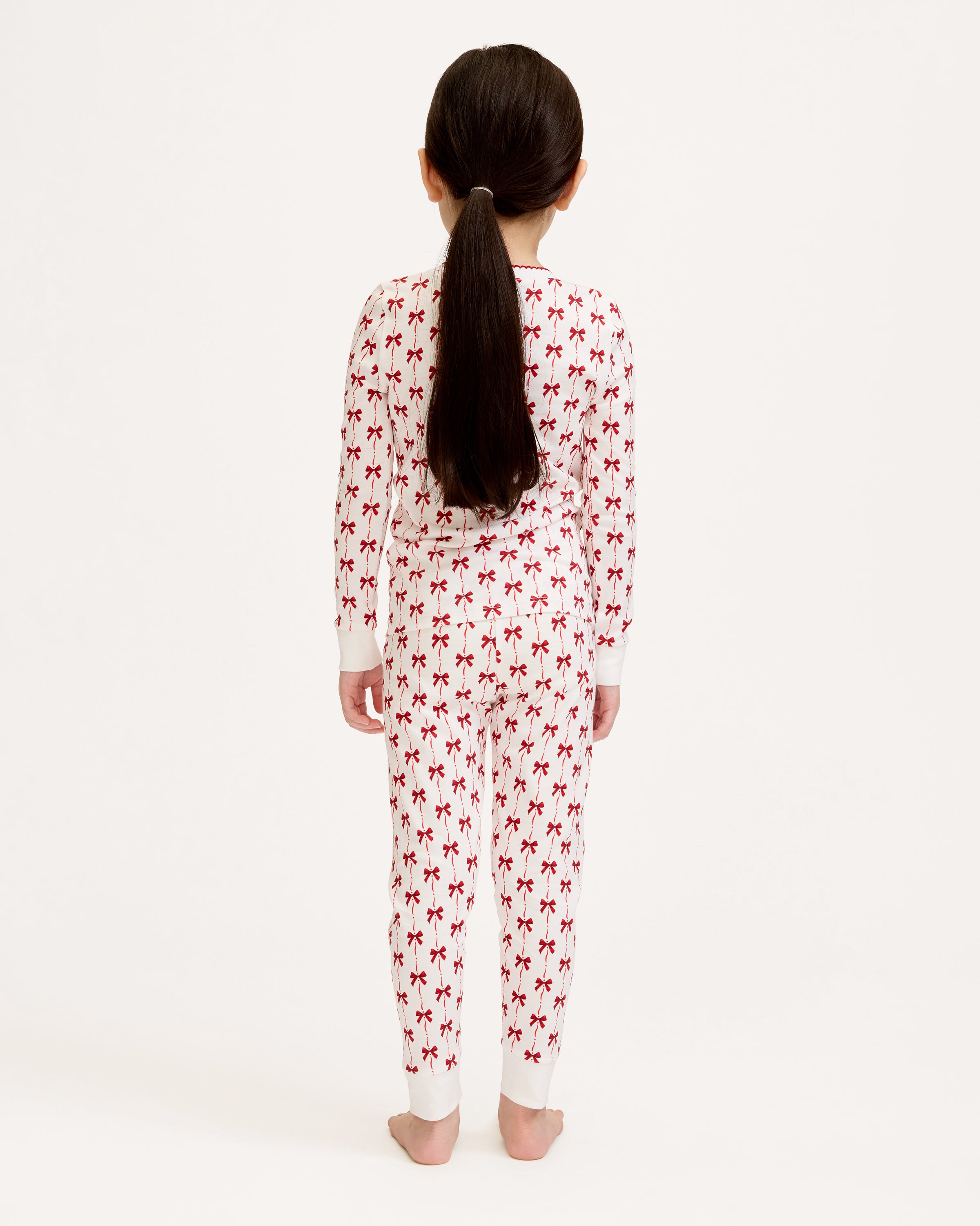 The Kid's Pima Snug Fit Pajama Set in Blissful Bows features a white two-piece design with long sleeves and pants, decorated with a repeating small red bow pattern and finished with white cuffs on the sleeves and pant legs.