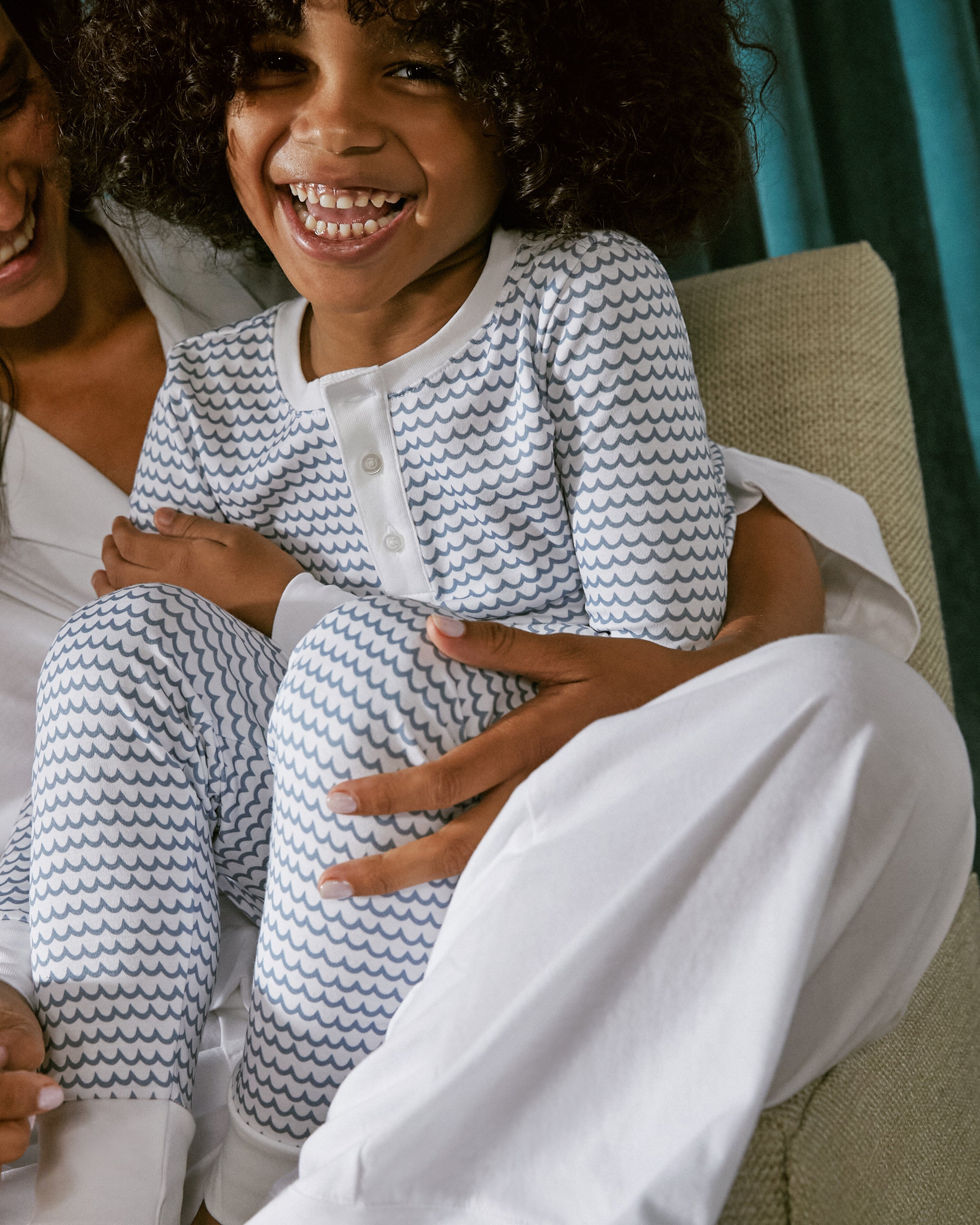 A curly-haired child sits smiling on an adults lap, both in white attire. The child wears a Petite Plume Kids Pima Snug Fit Pajama Set in La Mer, featuring wavy blue patterns. Teal curtains frame the cozy scene, enhancing its warmth.