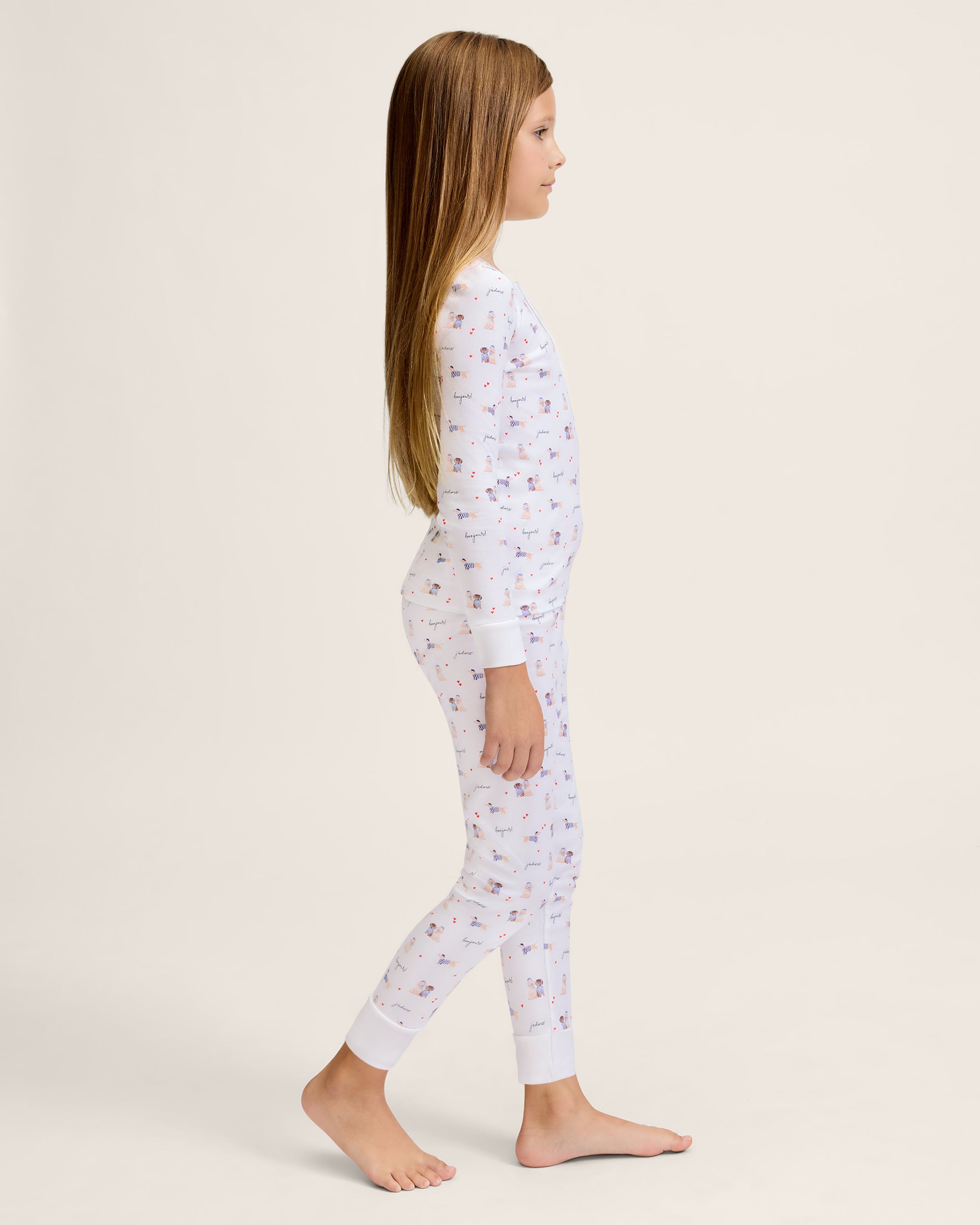 The Kid's Pima Snug Fit Pajama Set in Le Woof is a white long-sleeve pajama set with matching pants, featuring a colorful all-over cartoon dog and bone print, plus cuffed sleeves and ankles for a snug fit.