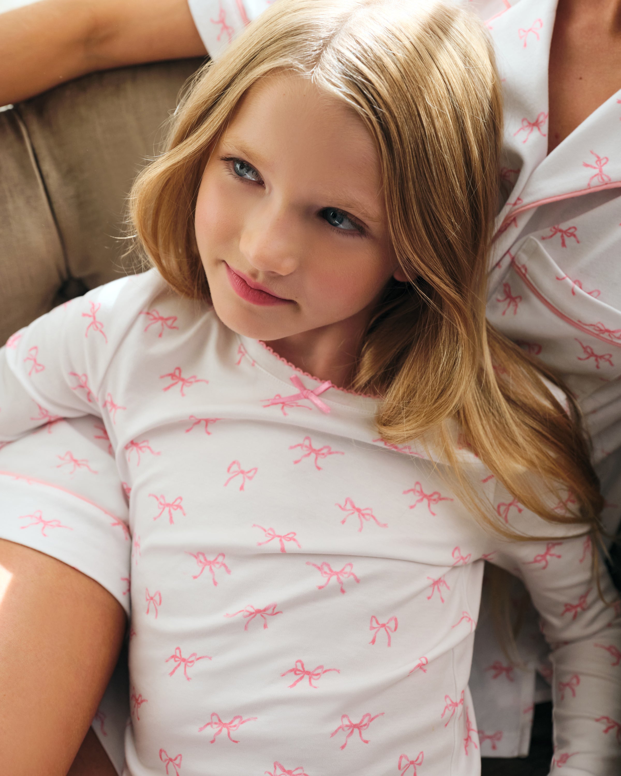 A confident young girl with long brown hair wears the Petite Plume Kids Pima Snug Fit Pajama Set in Blushing Bows, featuring a long sleeve top and pants made from soft Pima cotton with small pink patterns against a plain light-colored background.
