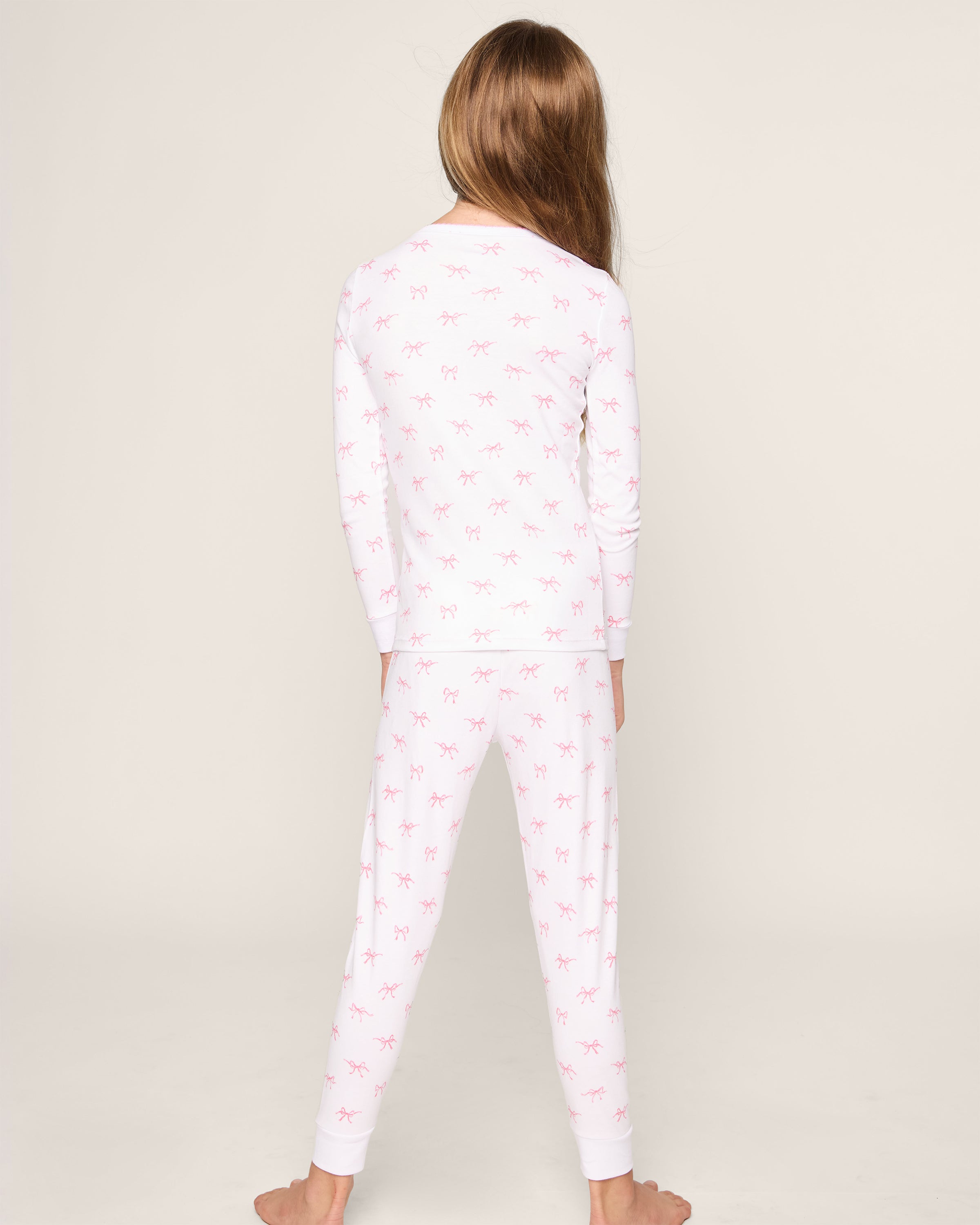 A child with long hair is seen from behind wearing Petite Plumes Kids Pima Snug Fit Pajama Set in Blushing Bows. The cozy sleepwear, made of soft Pima cotton and adorned with small pink bows, features a long sleeve top and pants. The child stands barefoot against a plain background.