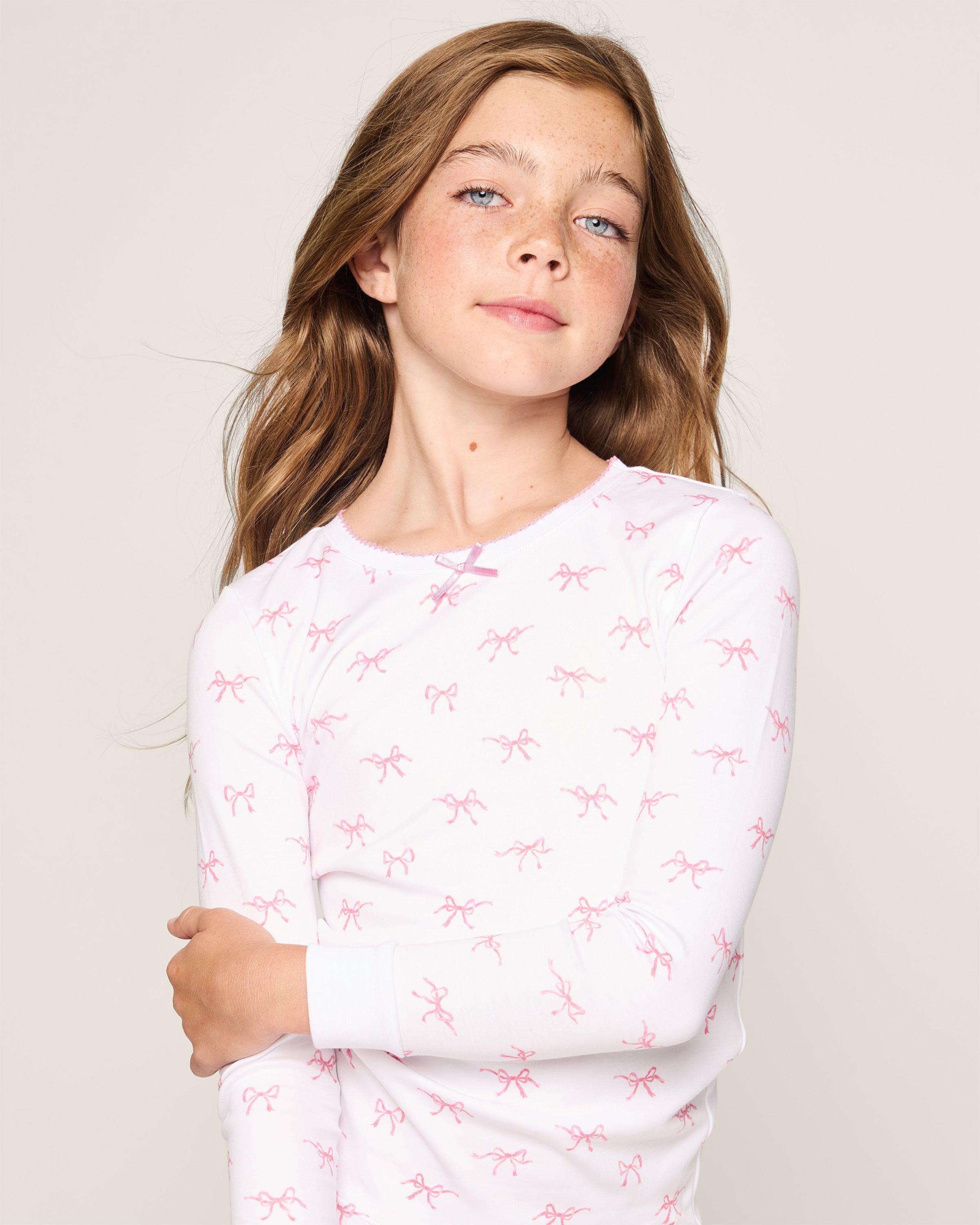 A young girl with long brown hair, wearing the Petite Plume Kids Pima Snug Fit Pajama Set in Blushing Bows, stands confidently against a plain background. Crafted from soft Pima cotton, her charming sleepwear features a white long-sleeved top with pink bow patterns as she gazes toward the camera.