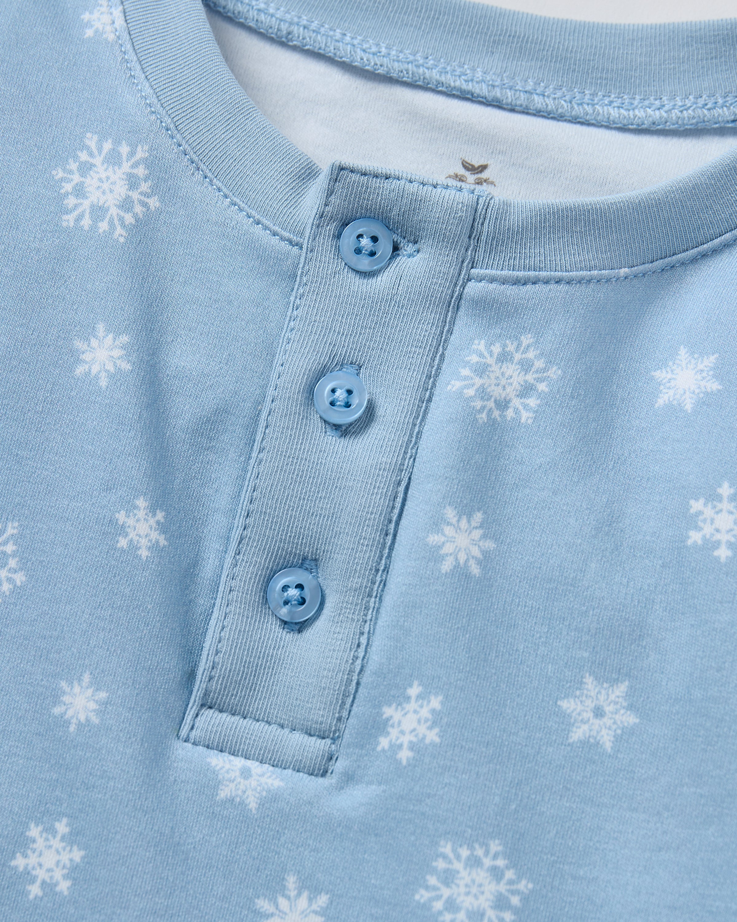 The Kid's Pima Snug Fit Pajama Set in Periwinkle Wonderland features light blue, long-sleeve pajamas with white snowflakes, buttoned neckline, matching elastic waist pants, and cuffed ankles. Shown laid flat on a white background.