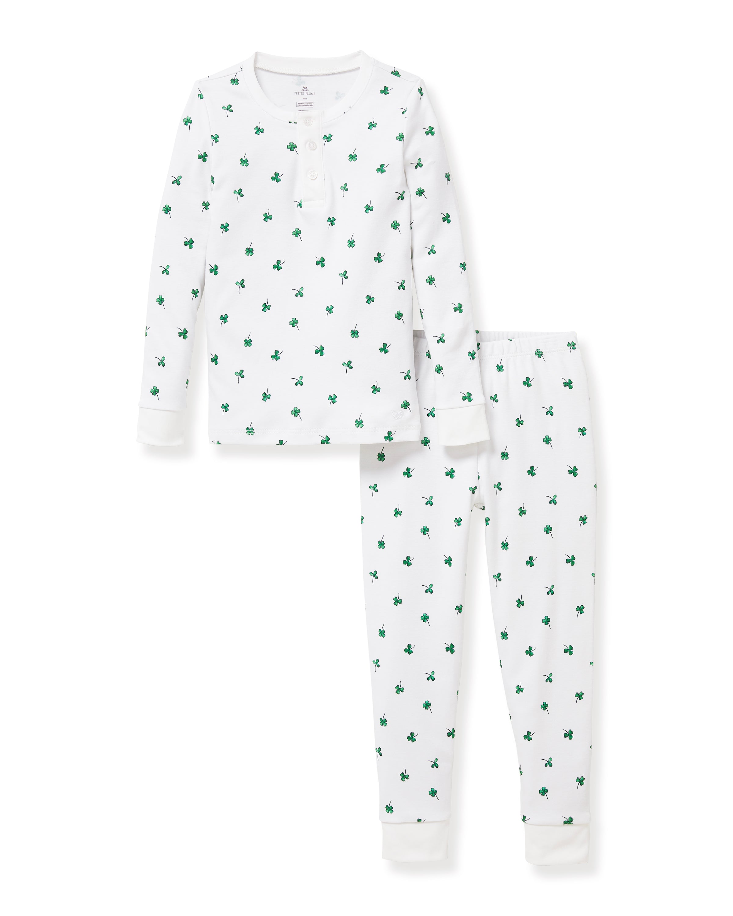 A young child with blonde hair smiles barefoot against a plain light background, dressed in the Kid's Pima Snug Fit Pajama Set in Shamrocks—white long-sleeve pajamas featuring small green shamrock patterns.