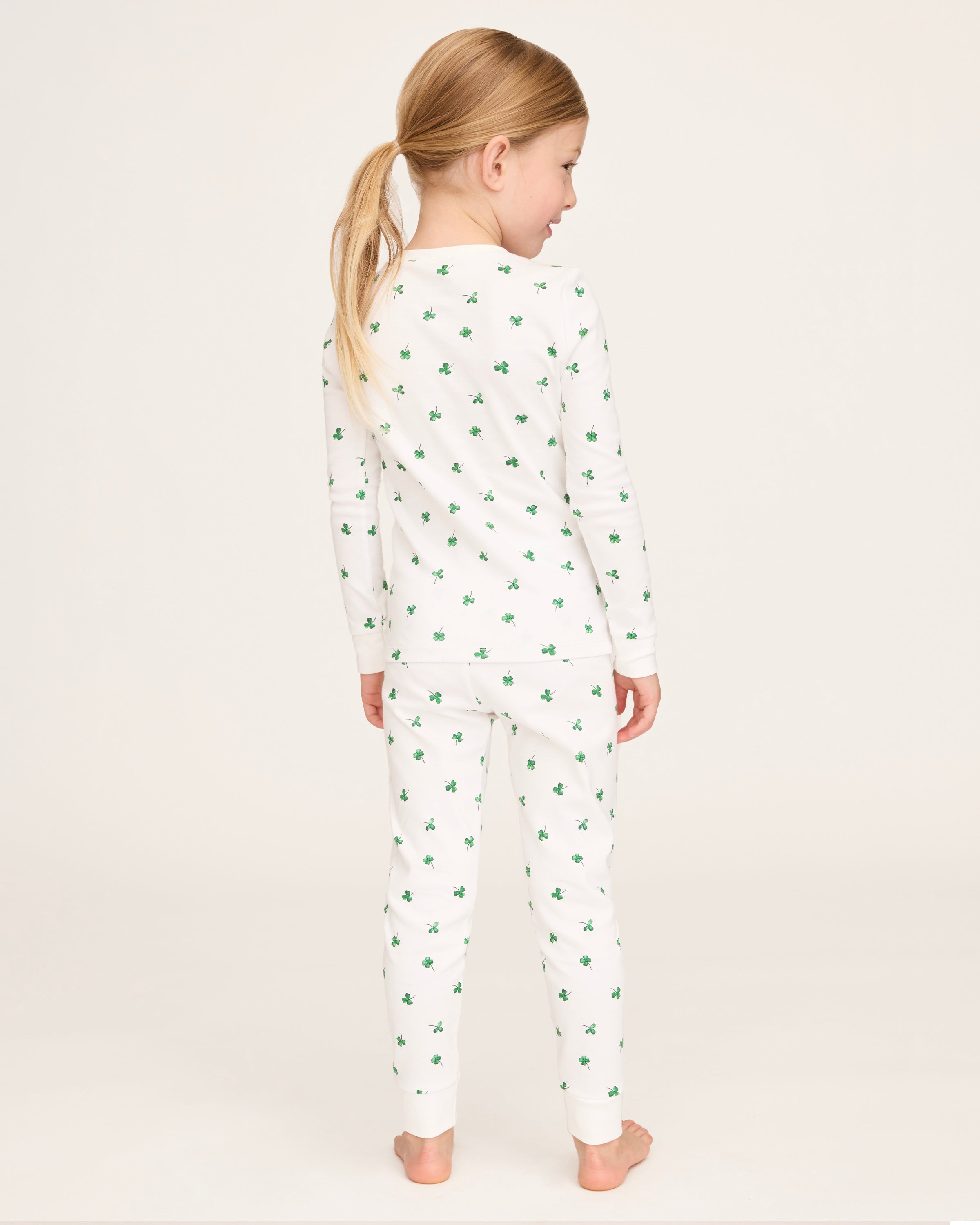 A young child with blonde hair smiles barefoot against a plain light background, dressed in the Kid's Pima Snug Fit Pajama Set in Shamrocks—white long-sleeve pajamas featuring small green shamrock patterns.