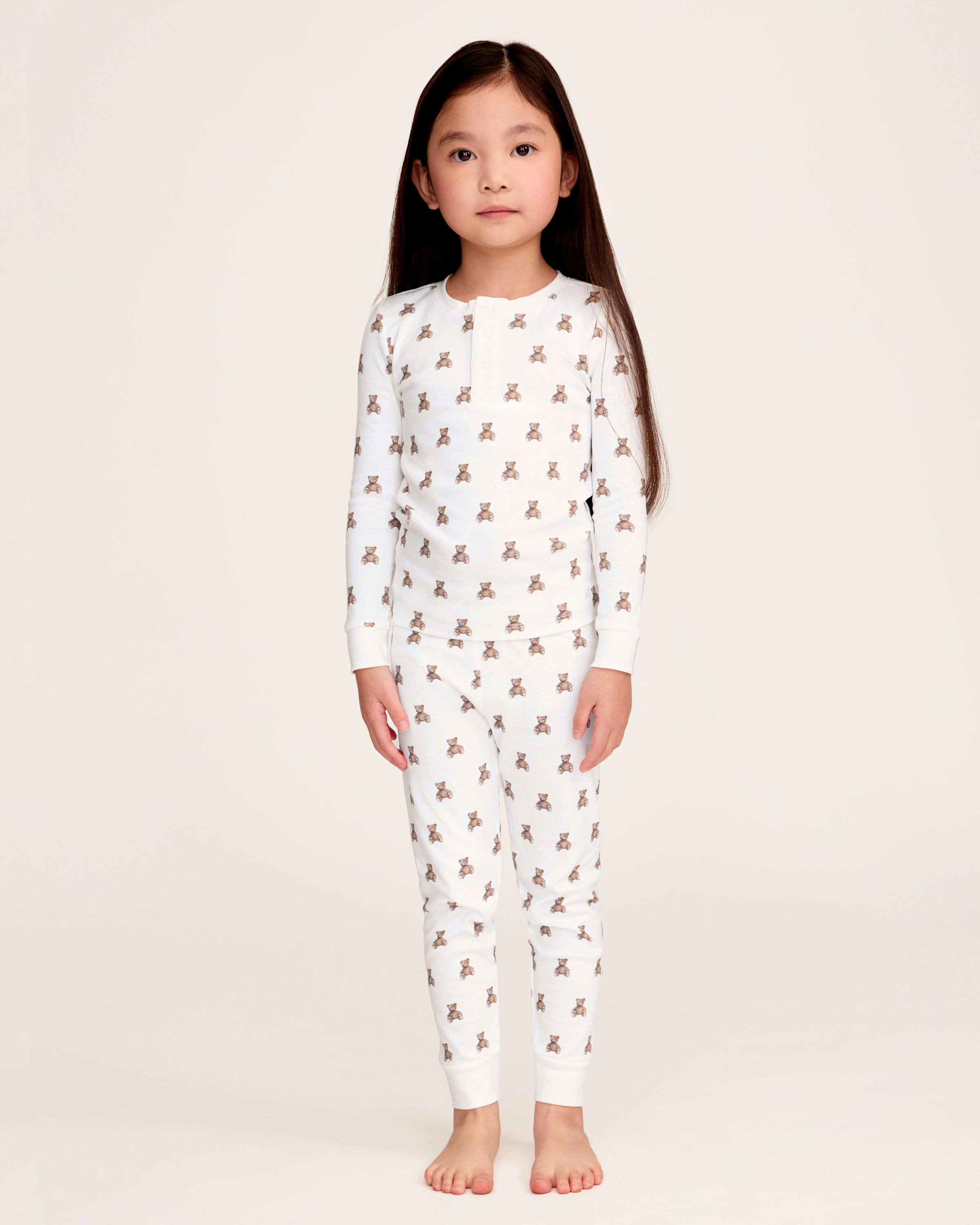 The Kid's Pima Snug Fit Pajama Set in Teddy Tales features a white long-sleeve buttoned top and elastic waist pants with ribbed cuffs, both printed all over with brown teddy bears. Suitable for children.