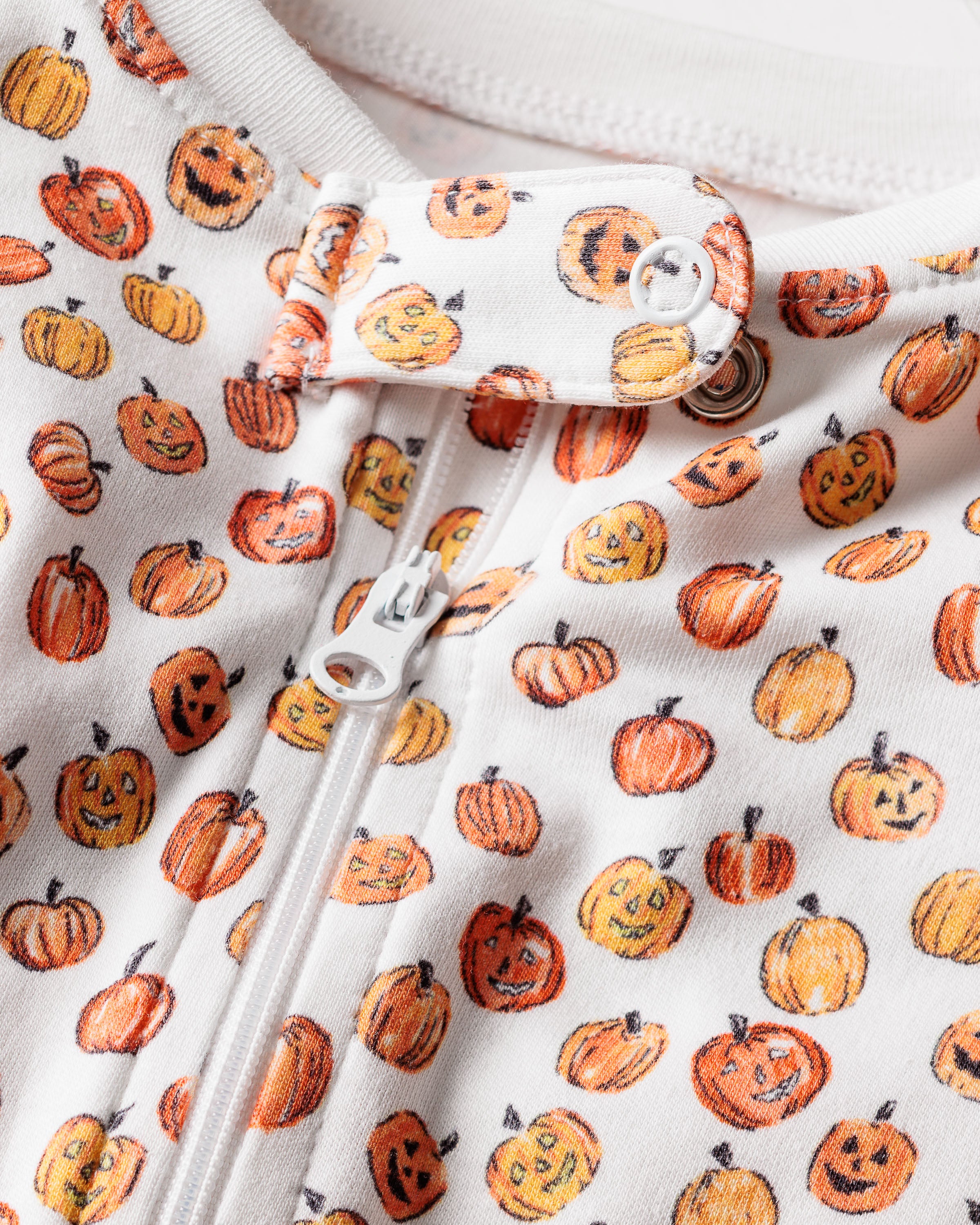 Close-up of a zipper on the white Pima cotton fabric of the Babys Pima Romper in Pumpkin Patch by Petite Plume, displaying orange pumpkins and Jack-o-lanterns with various expressions. This festive piece is ideal for cozy Halloween attire as kids pajamas or a romper.