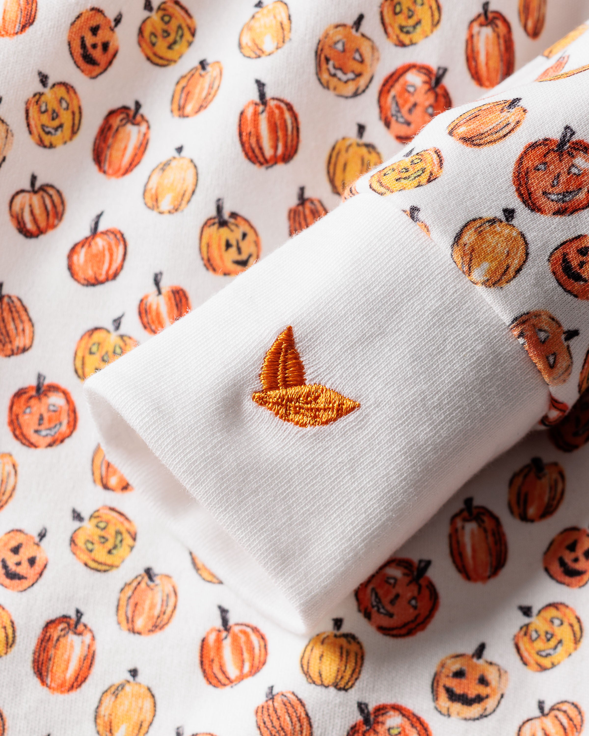 Close-up of Babys Pima Romper in Pumpkin Patch by Petite Plume, featuring a white romper with orange jack-o-lanterns in various sizes and expressions. The soft Pima cotton and embroidered orange leaf on the sleeve cuff make it perfect Halloween attire for kids.