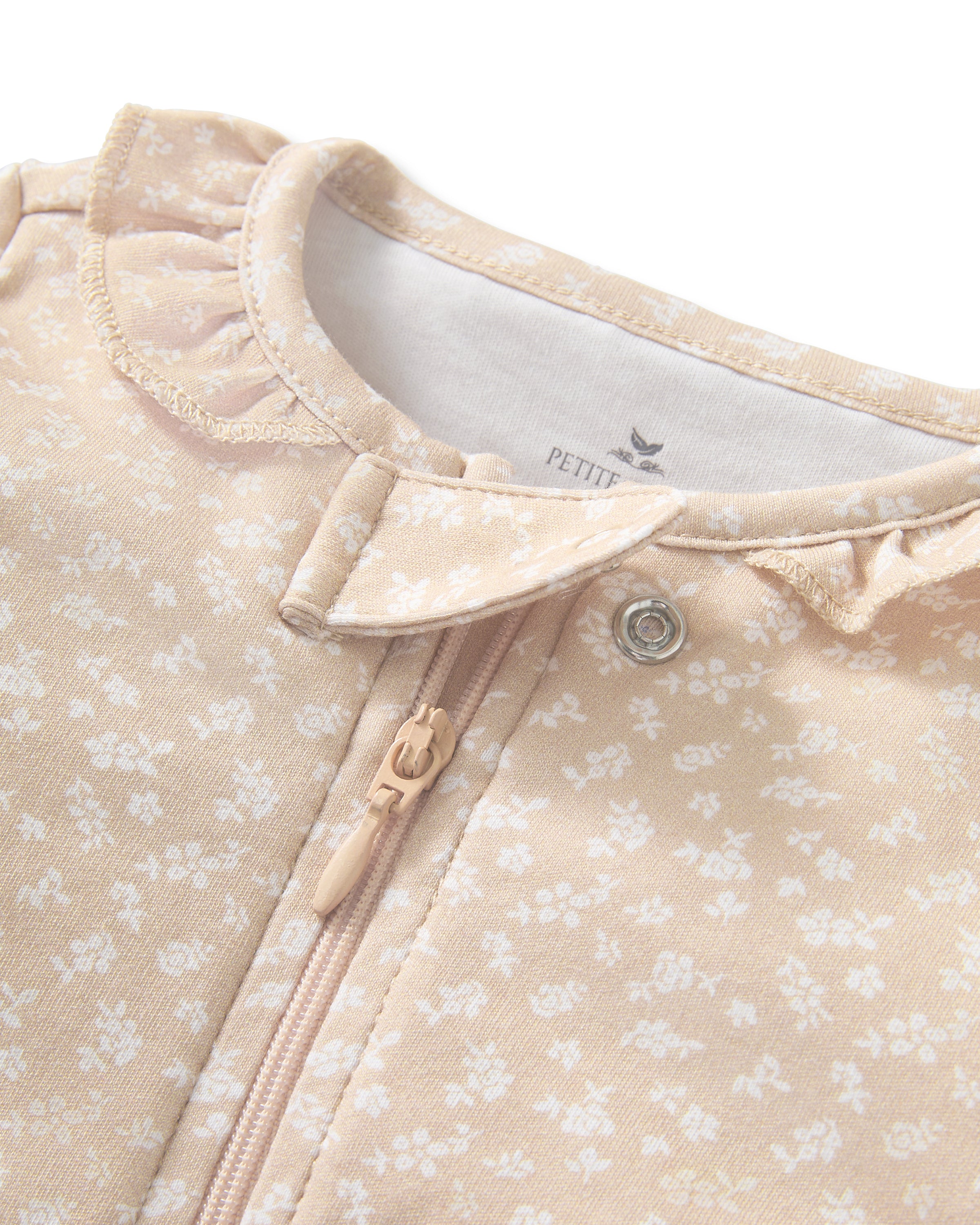 The Baby's Pima Romper in Ditsy Waltz Camel features a beige color with a white floral pattern, long sleeves, covered feet, fold-over cuffs, a ruffled collar, and a diagonal zipper closure.