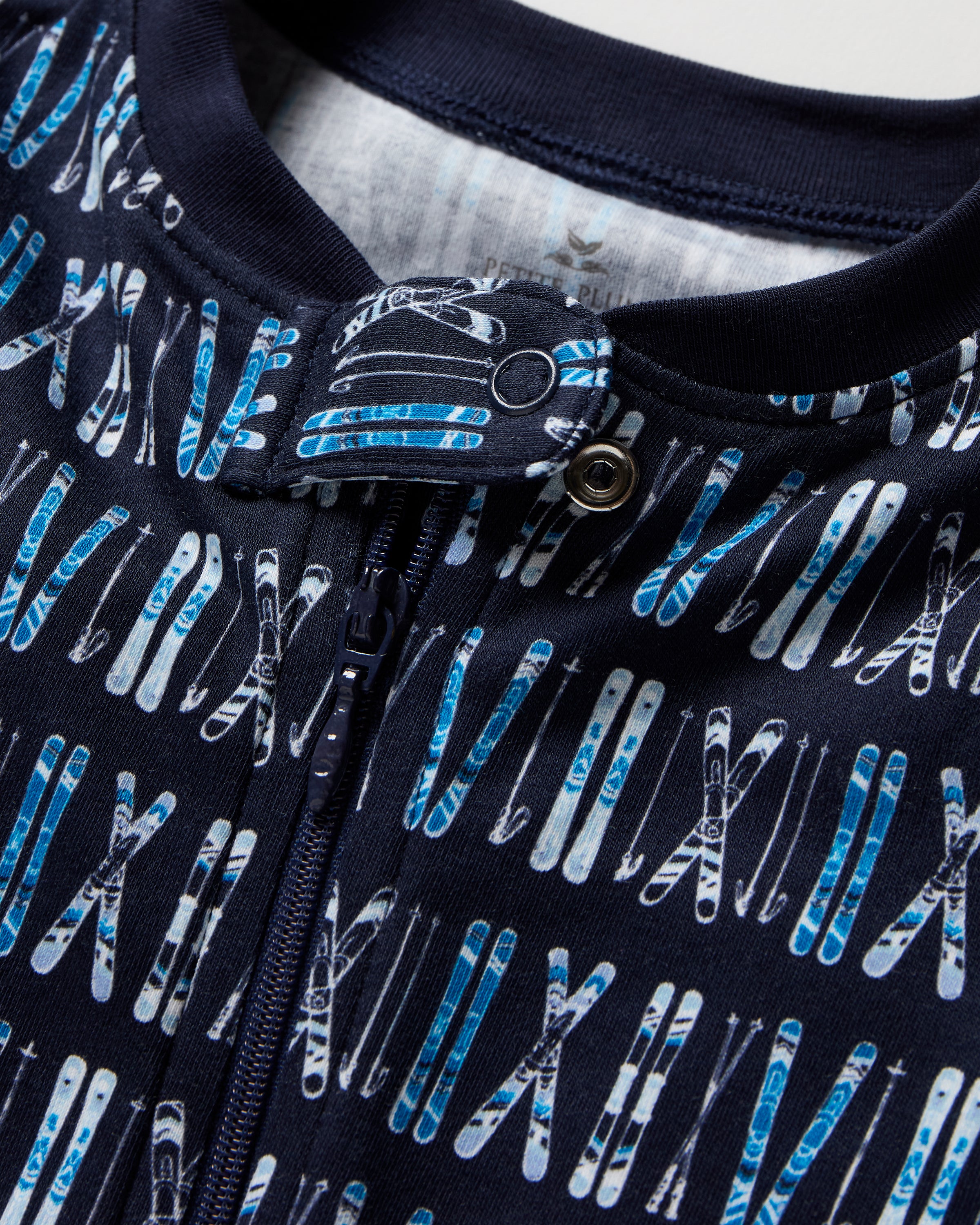 The Baby's Pima Romper in Ski Trails is a navy blue zip-up footed onesie with an all-over pattern of white and blue skis and ski poles arranged vertically.