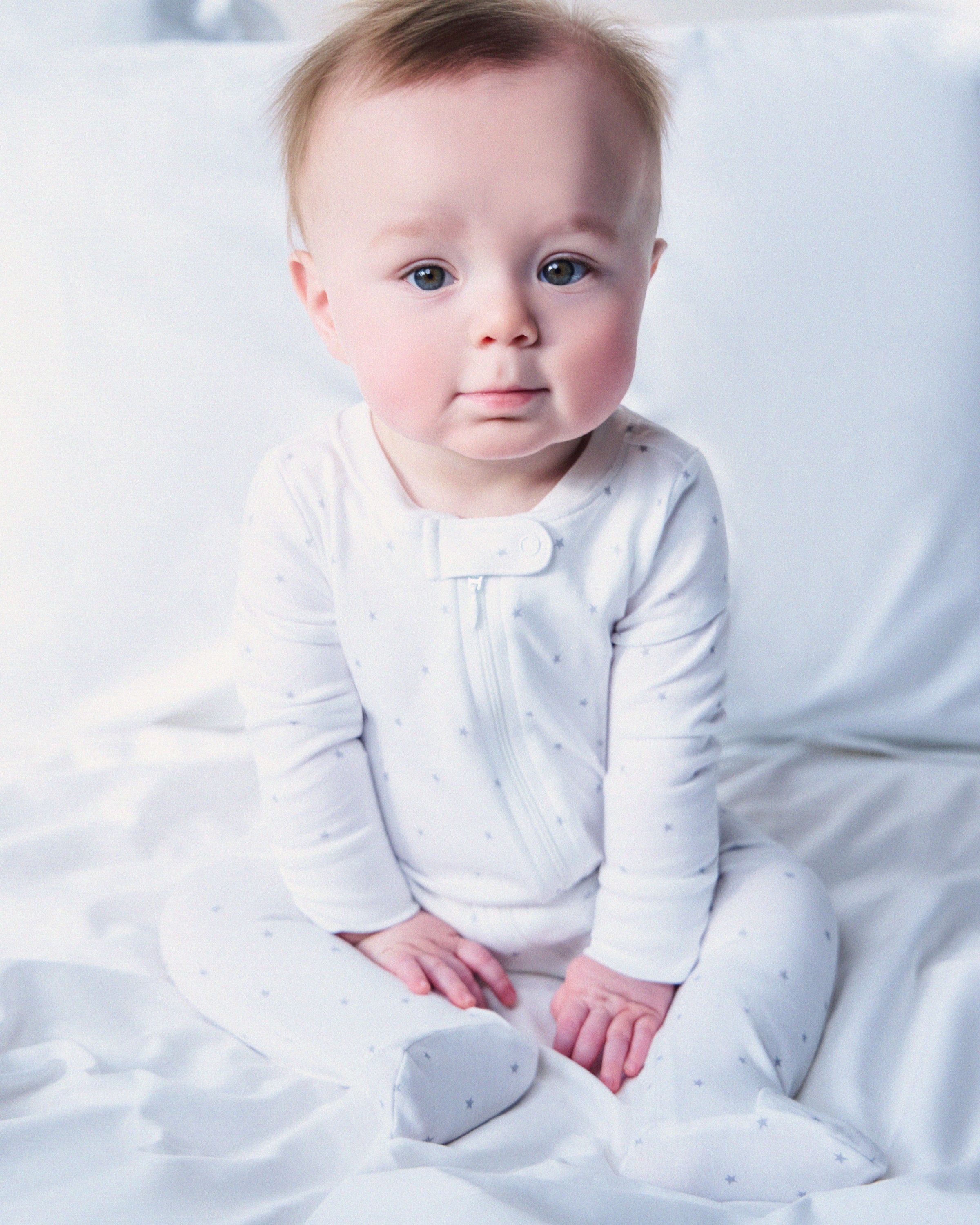 A baby sits on white sheets in a luxurious Baby’s Pima Romper in Grey Stars by Petite Plume, crafted from soft Pima cotton. The romper features a subtle pattern and a front zipper. The baby, with light hair and a calm expression, gazes slightly to the side.