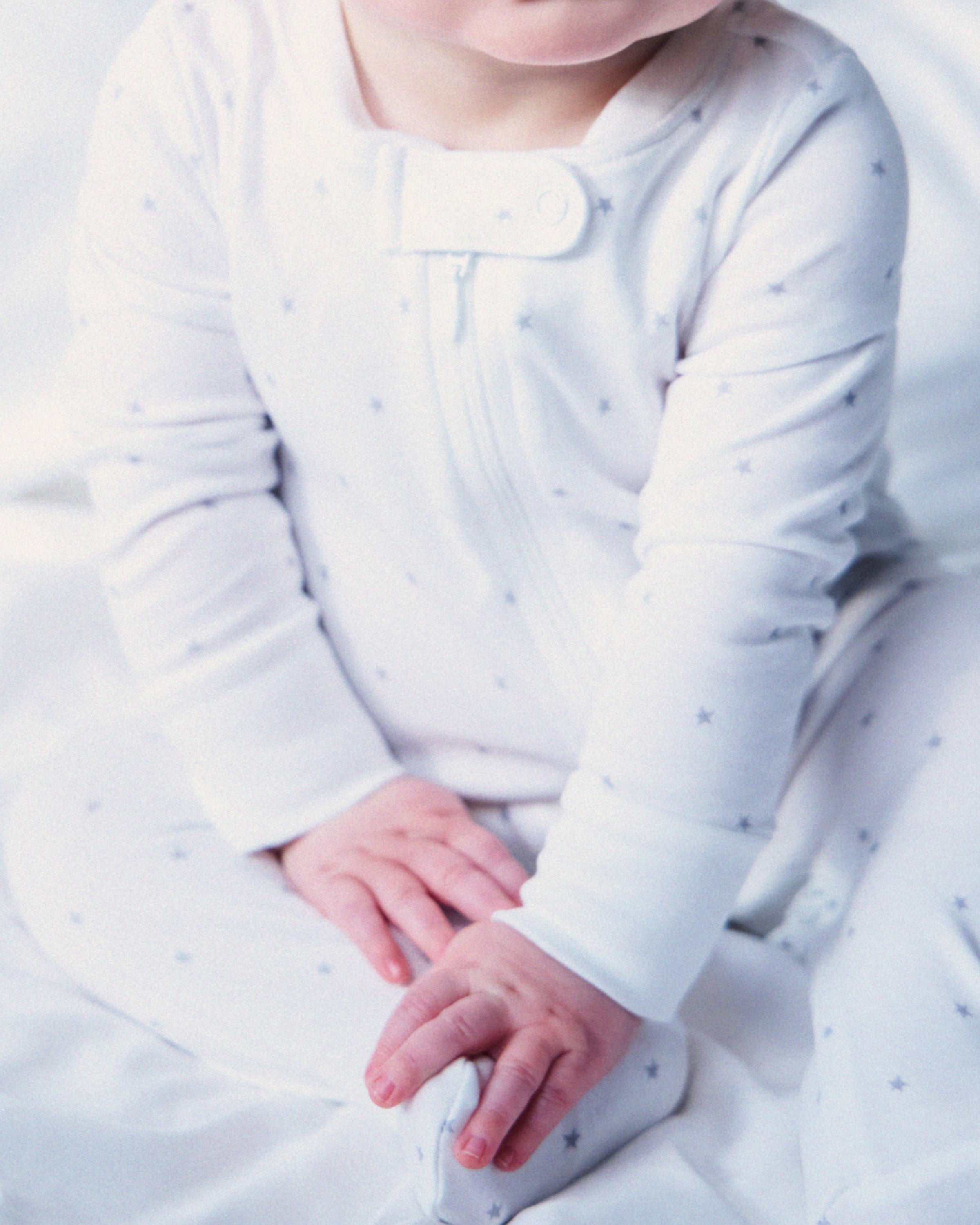 A baby in Petite Plumes Babys Pima Romper in Grey Stars sits on a soft white surface. Their hands rest gently on their legs, showcasing the rompers elegance, with the face partially out of view.
