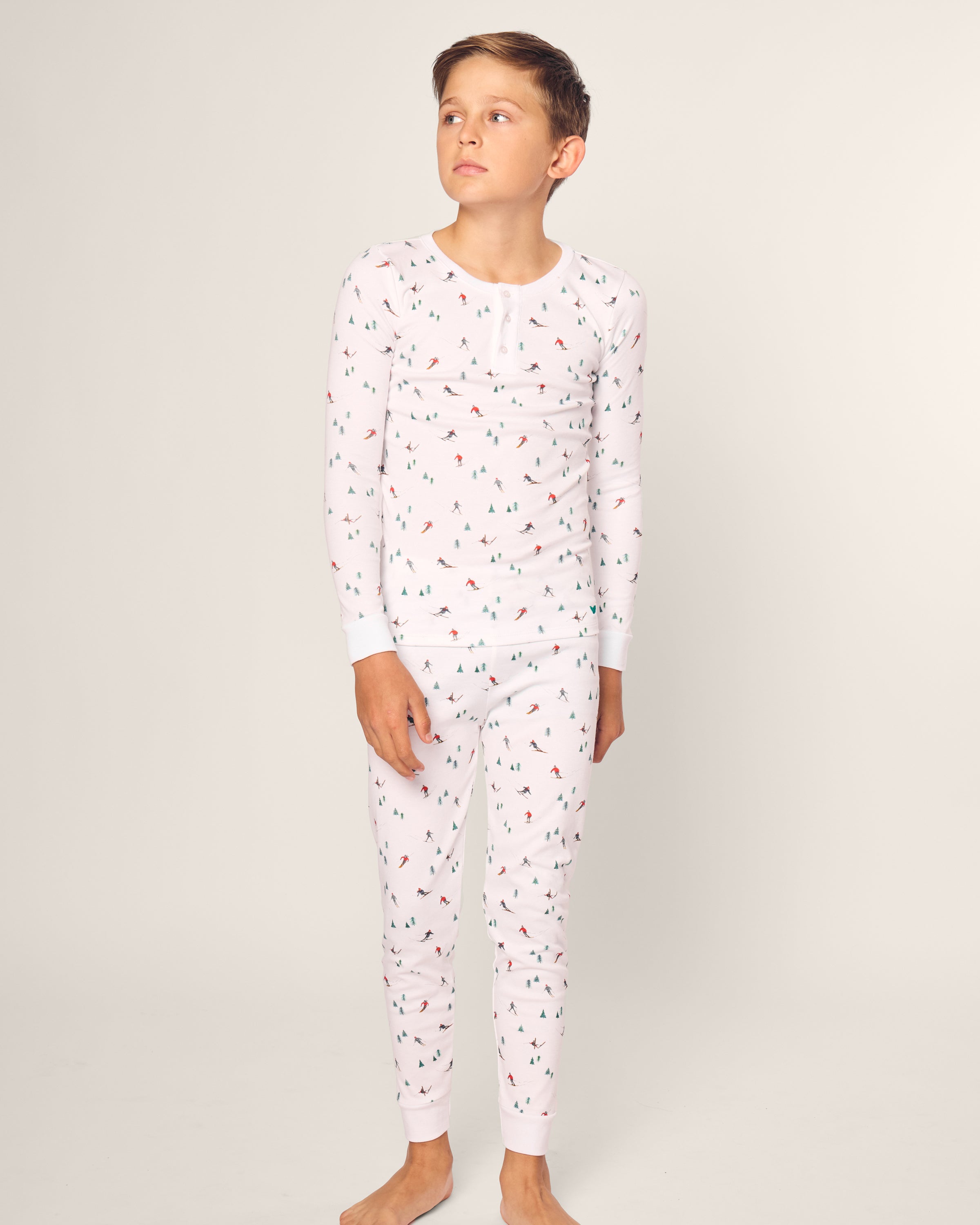 A child stands barefoot against a plain background, wearing Petite Plumes Kids Pima Snug Fit Pajama Set in Après Ski. The snug long sleeve top features a playful pattern with small trees, tents, and campfires, crafted from soft Pima cotton with a classic button-up style.