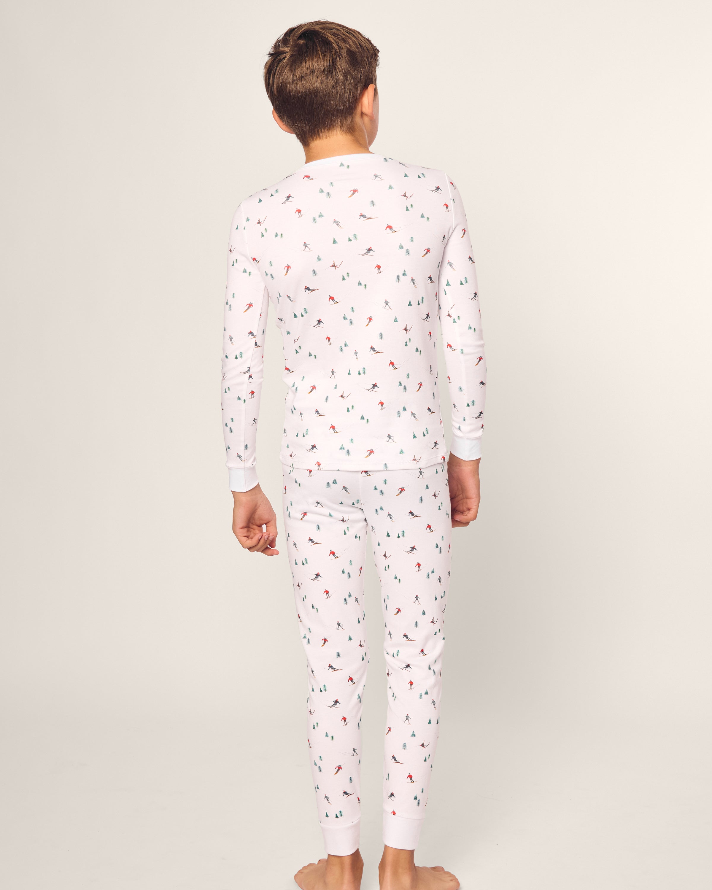 A child stands barefoot, facing away in a snug-fitting Petite Plume Kids Pima Snug Fit Pajama Set in Après Ski. Made from soft Pima cotton with colorful prints, the pajamas feature long sleeves and pants. The background is a plain, light color.